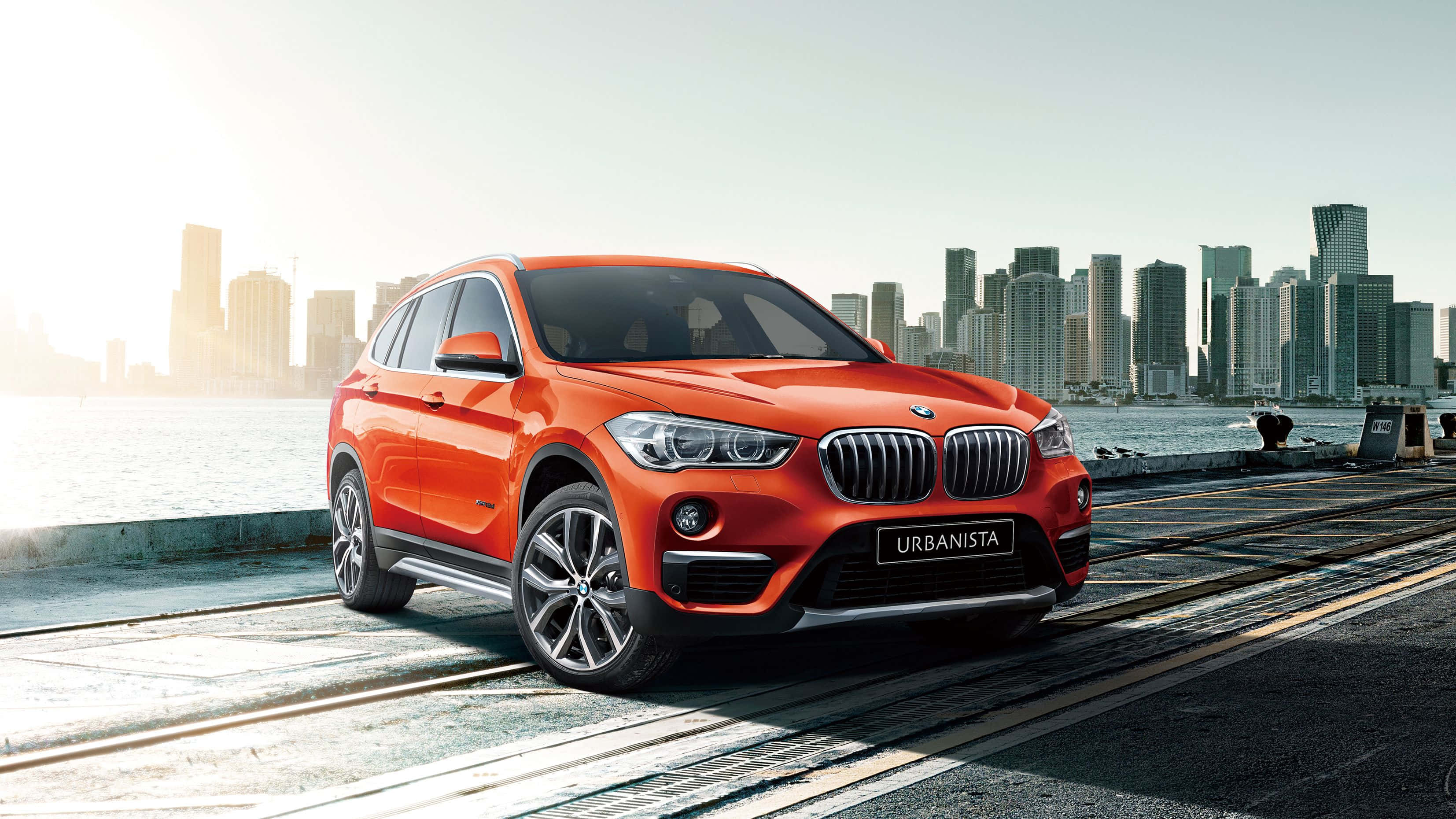 Bmw X1 桌布 壁紙 — 一輛鮮橙色的BMW X1 Urbanista SUV停在濱海長廊上，背景是現代化的城市天際線。
