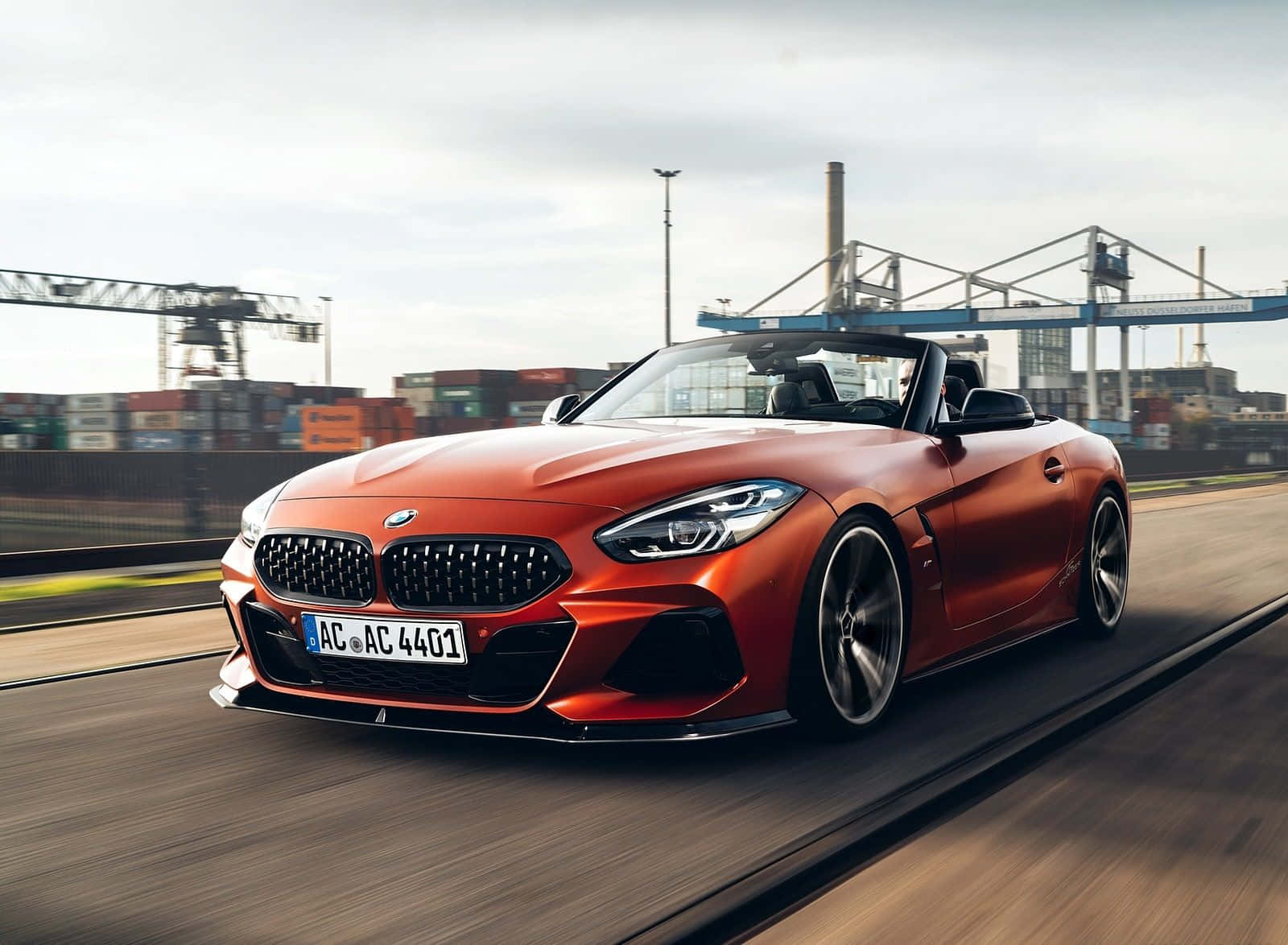 Bmw Z4 桌布 壁紙 — 一輛鮮橙色的BMW Z4敞篷跑車在高速公路上疾馳，背景模糊中可見工業起重機和貨運貨櫃。