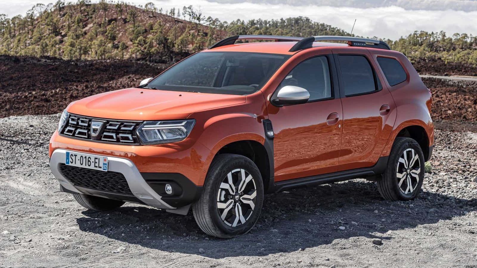 Dacia 桌布 壁紙 — 一輛鮮橙色的Dacia Duster SUV停放在碎石上，背景是陽光明媚下的森林山丘。