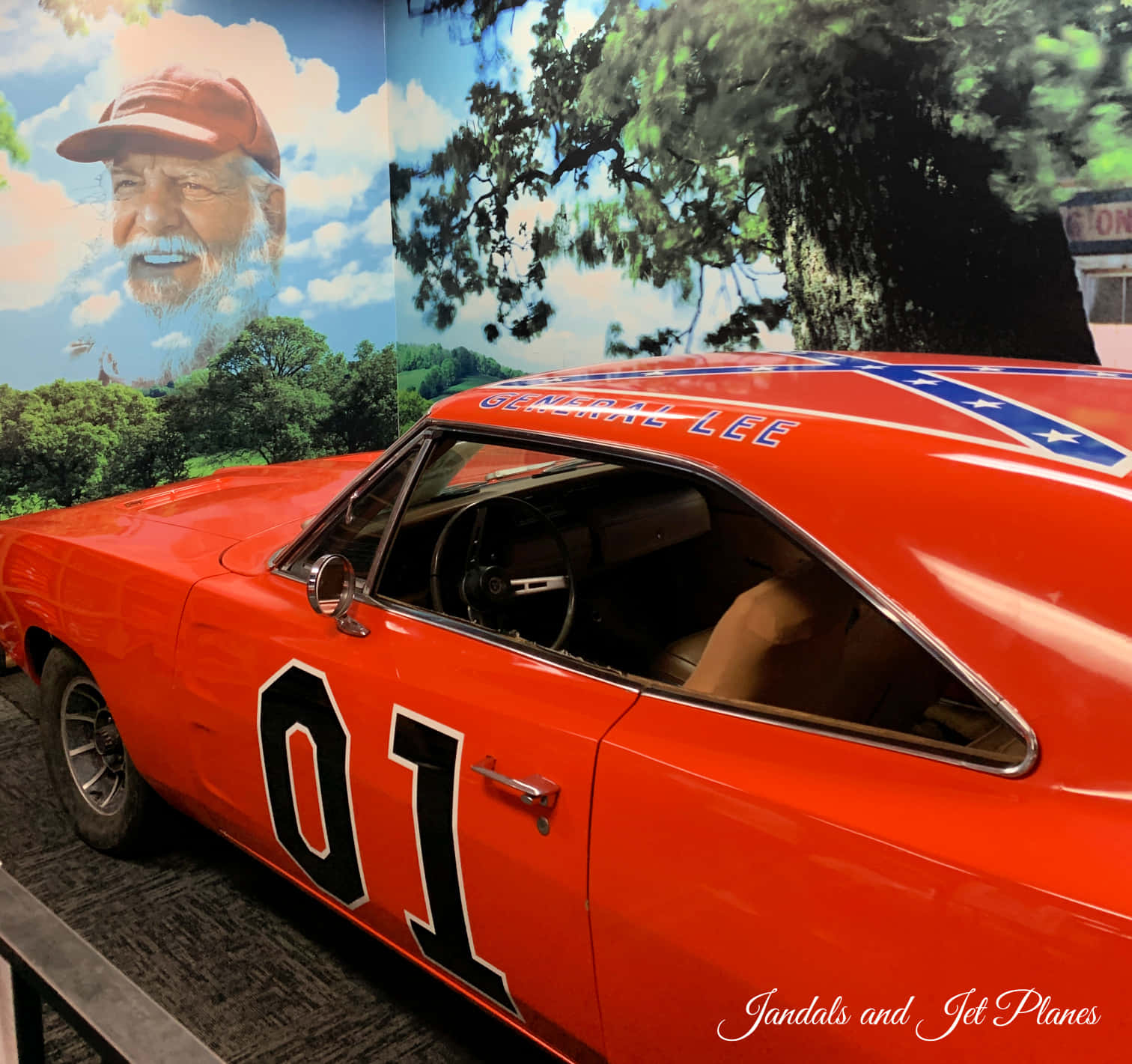 General Lee 車 桌布 壁紙 — 一輛鮮橙色的General Lee車，帶有'01'和聯邦旗貼紙，展示在一個鬍鬚男人和樹木的壁畫前面。
