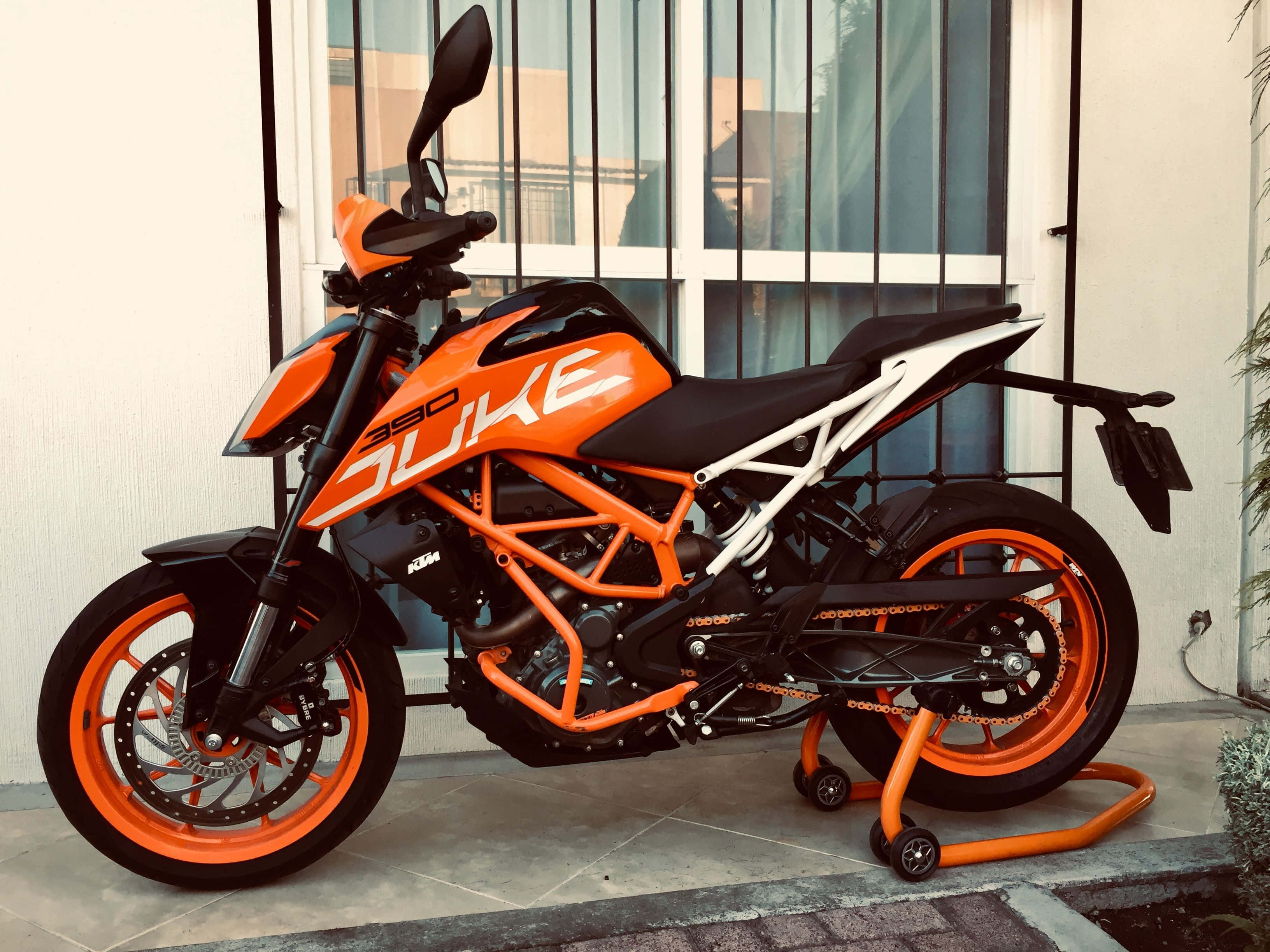 橘色 桌布 壁紙 — 一輛鮮橙色的KTM 390 Duke機車停放在磁磚庭院上，後面有一座現代建築。