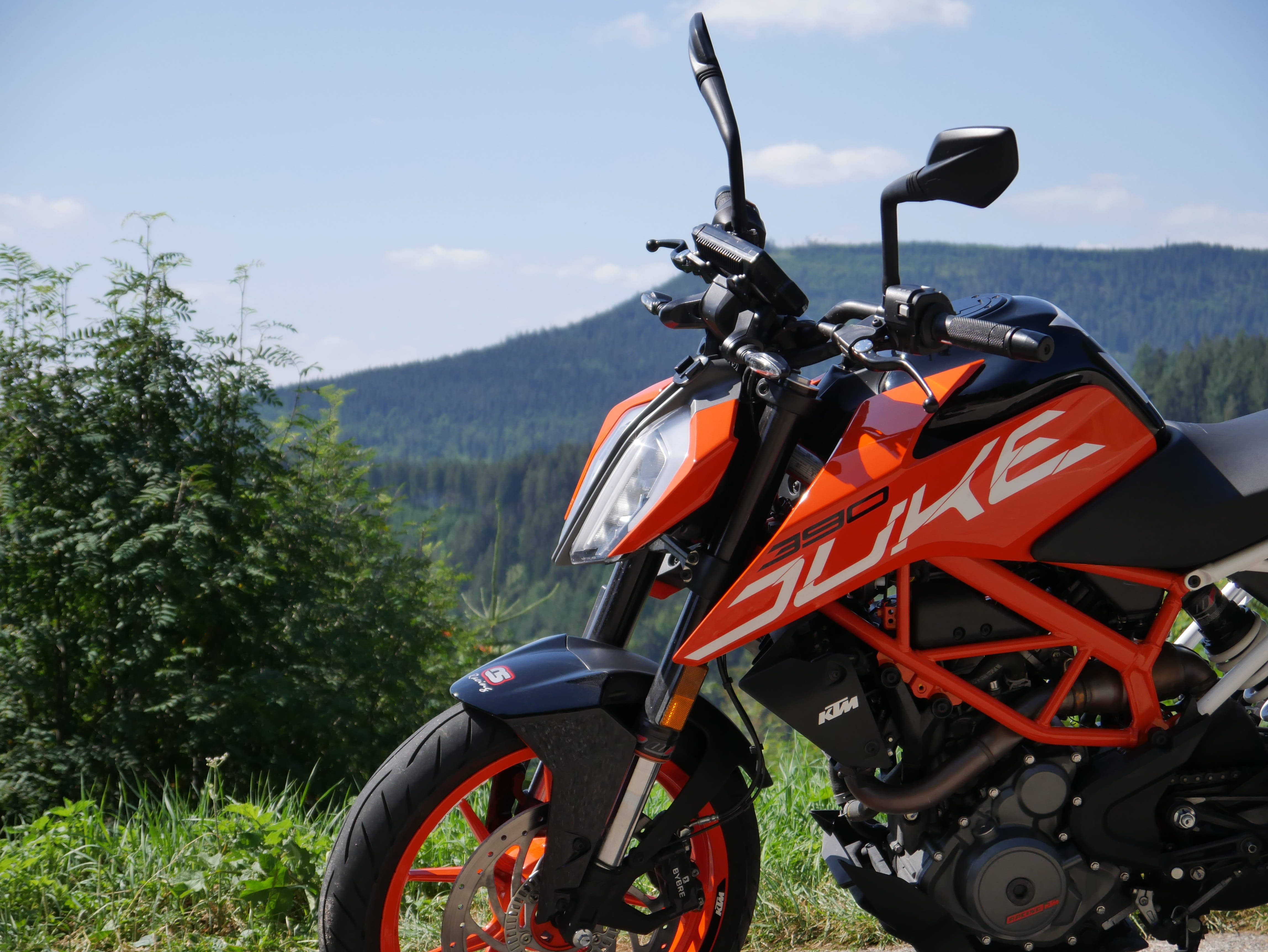 KTM 摩托車 桌布 壁紙 — 一輛鮮橙色的KTM Duke 390機車停放在一個草地山坡上，背景是森林覆蓋的山脈，天空清澈湛藍。