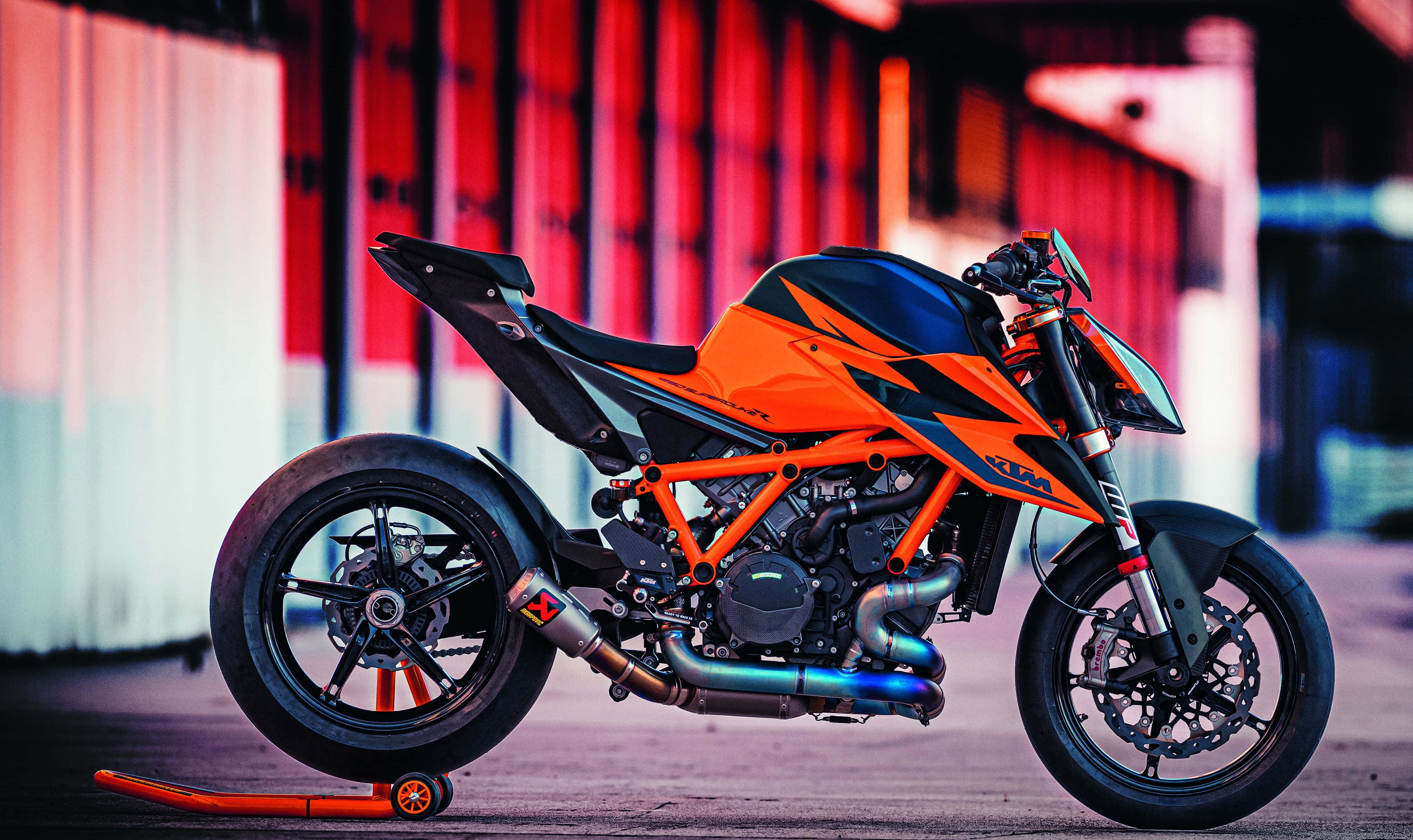 Ktm Bike 桌布 壁紙 — 一輛鮮橙色的KTM摩托車停在支架上，背景是模糊的紅白工業場景。