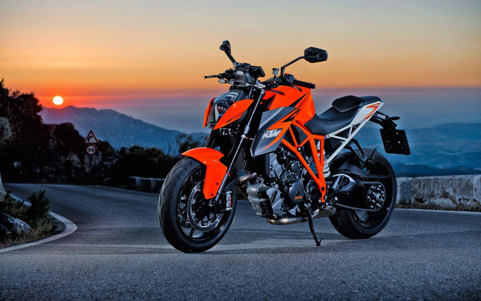 KTM 摩托車 桌布 壁紙 — 一輛鮮橙色的KTM機車停泊在蜿蜒的山路上，夕陽在遠山後發出光芒。
