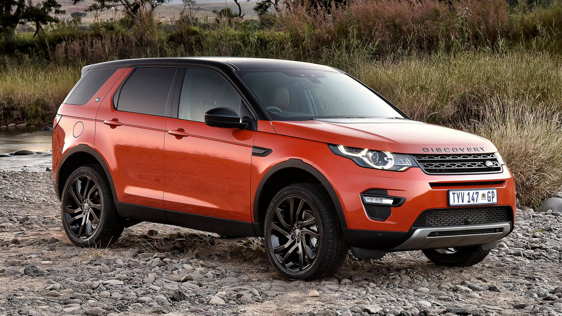 land rover iPhone 桌布 壁紙 — 一輛鮮橙色的Land Rover Discovery停泊在岩石河岸上，背景有高草和樹木。