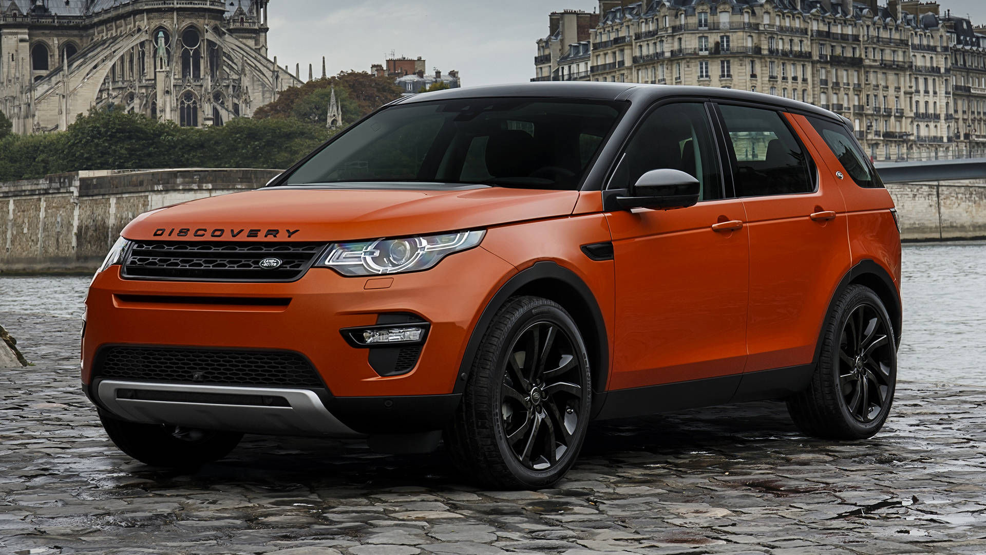 land rover iPhone 桌布 壁紙 — 一輛鮮橙色的Land Rover Discovery SUV停放在濕漉漉的鵝卵石路上，背景是巴黎聖母院和巴黎建築。