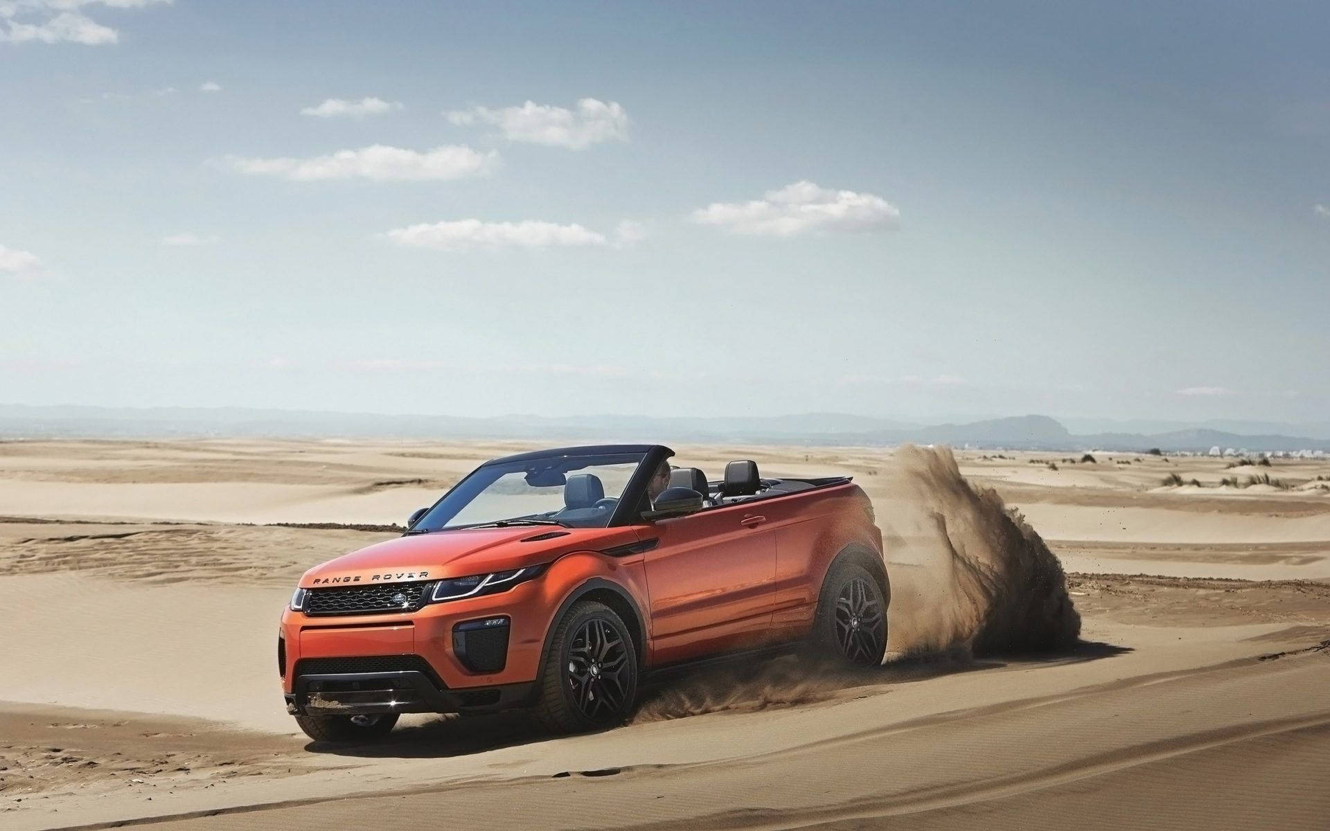 荒原路華 桌布 壁紙 — 一輛鮮橙色的Range Rover Evoque Convertible在晴朗的藍天下，飛馳過陽光照耀的沙漠沙丘，揚起一片沙塵。