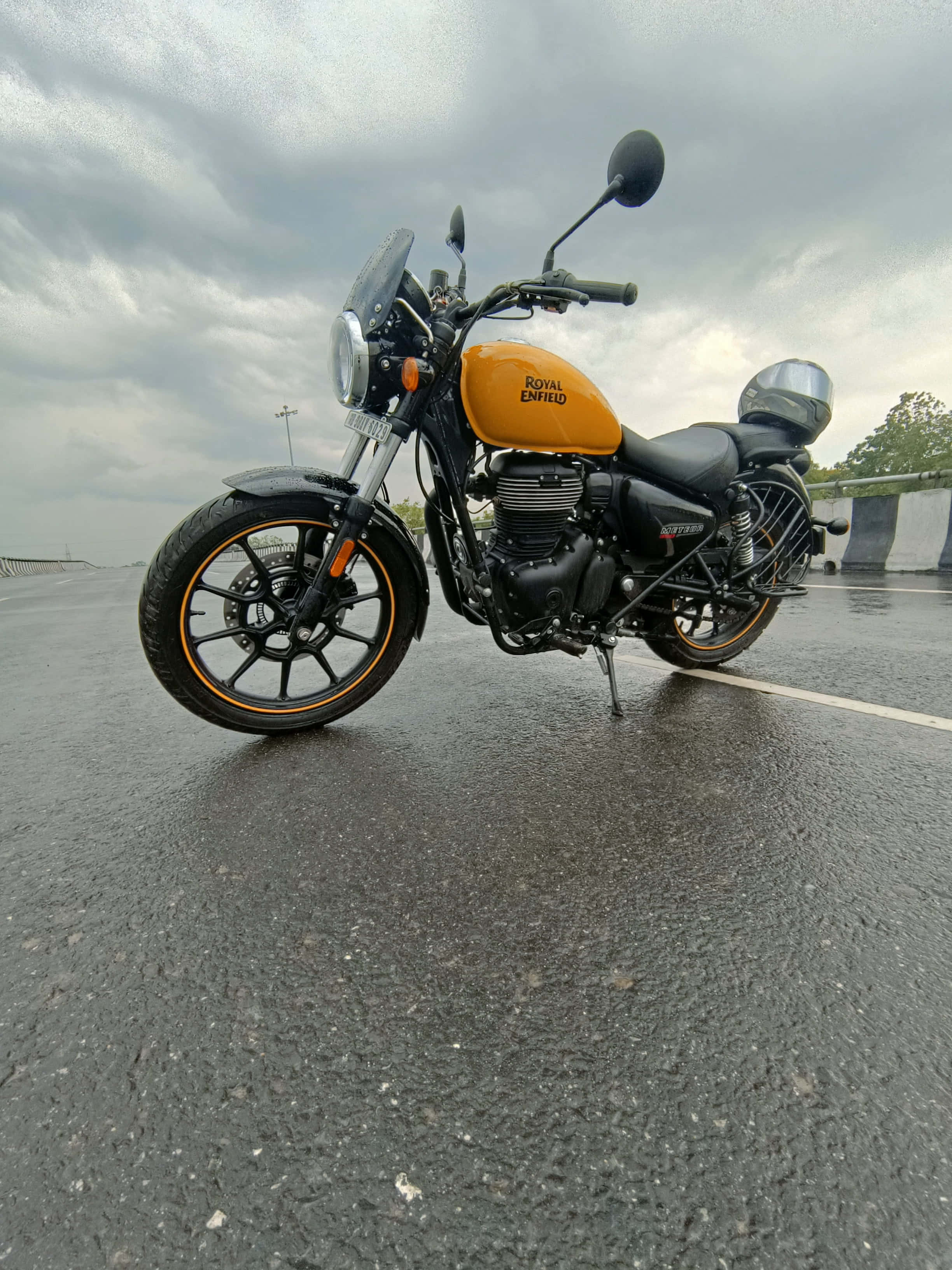 Royal Enfield Bullet 桌布 壁紙 — 一輛鮮橙色的Royal Enfield機車停在潮濕的柏油路上，遠處有樹木，天空多雲。