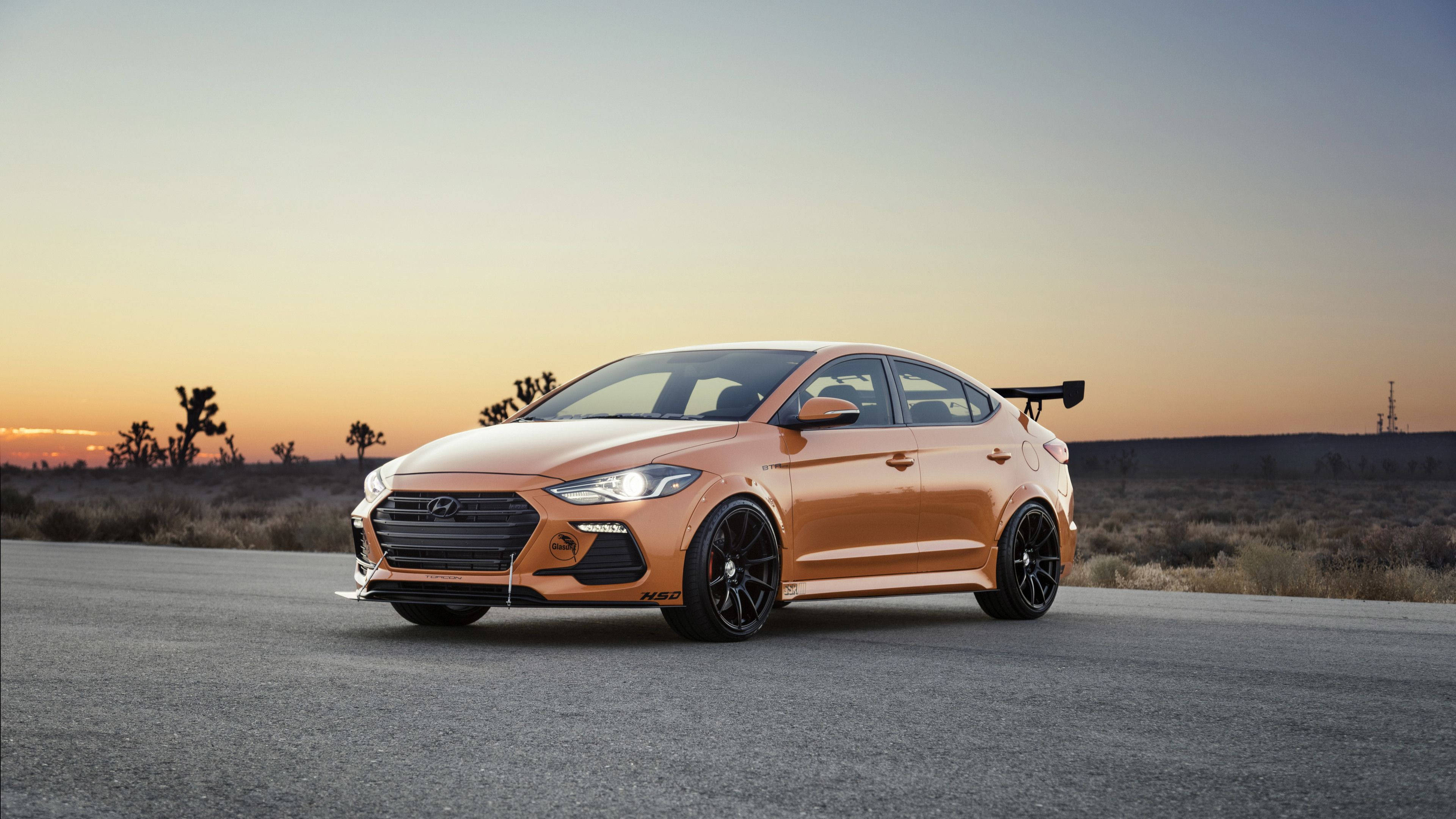Hyundai 桌布 壁紙 — 一輛鮮橙色的現代 Elantra N 停泊在沙漠公路上，背景是日落和約書亞樹。