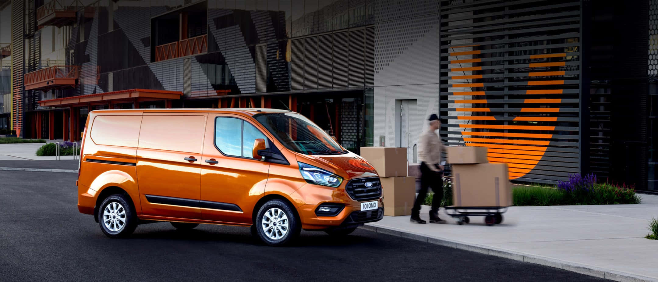 Ford Transit 桌布 壁紙 — 一輛鮮橙色的福特全順貨車停在一棟現代建築旁，一名工人推著裝滿紙箱的手推車。