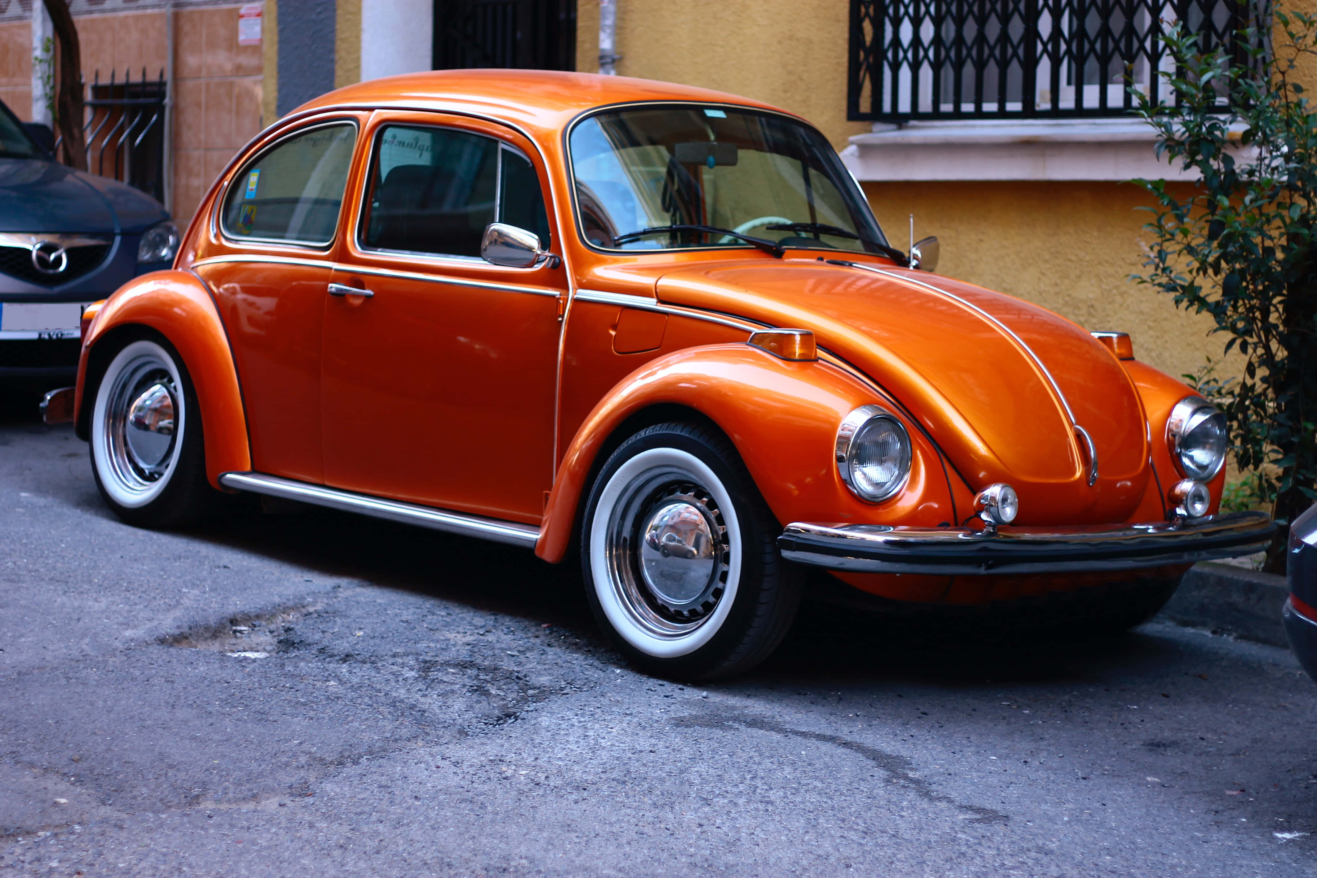 Vintage Car 桌布 壁紙 — 一輛鮮橙色的經典福斯金龜車停泊在陽光普照的市區街道上，旁邊是一座黃色建築物。