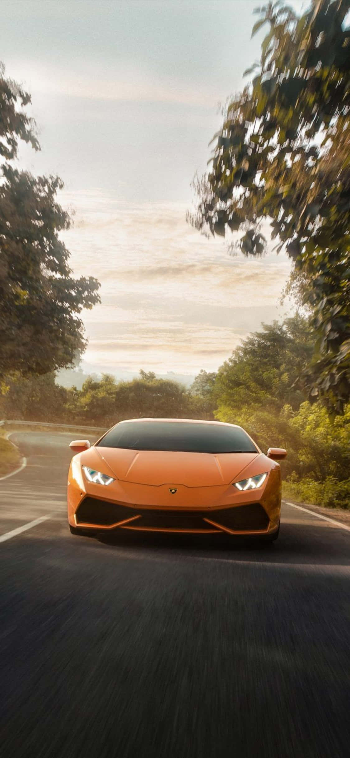 iPhone X Lamborghini 桌布 壁紙 — 一輛鮮橙色的藍寶堅尼 Huracán 在黃金時段沿著蜿蜒的森林道路高速行駛，帶有動態模糊效果。