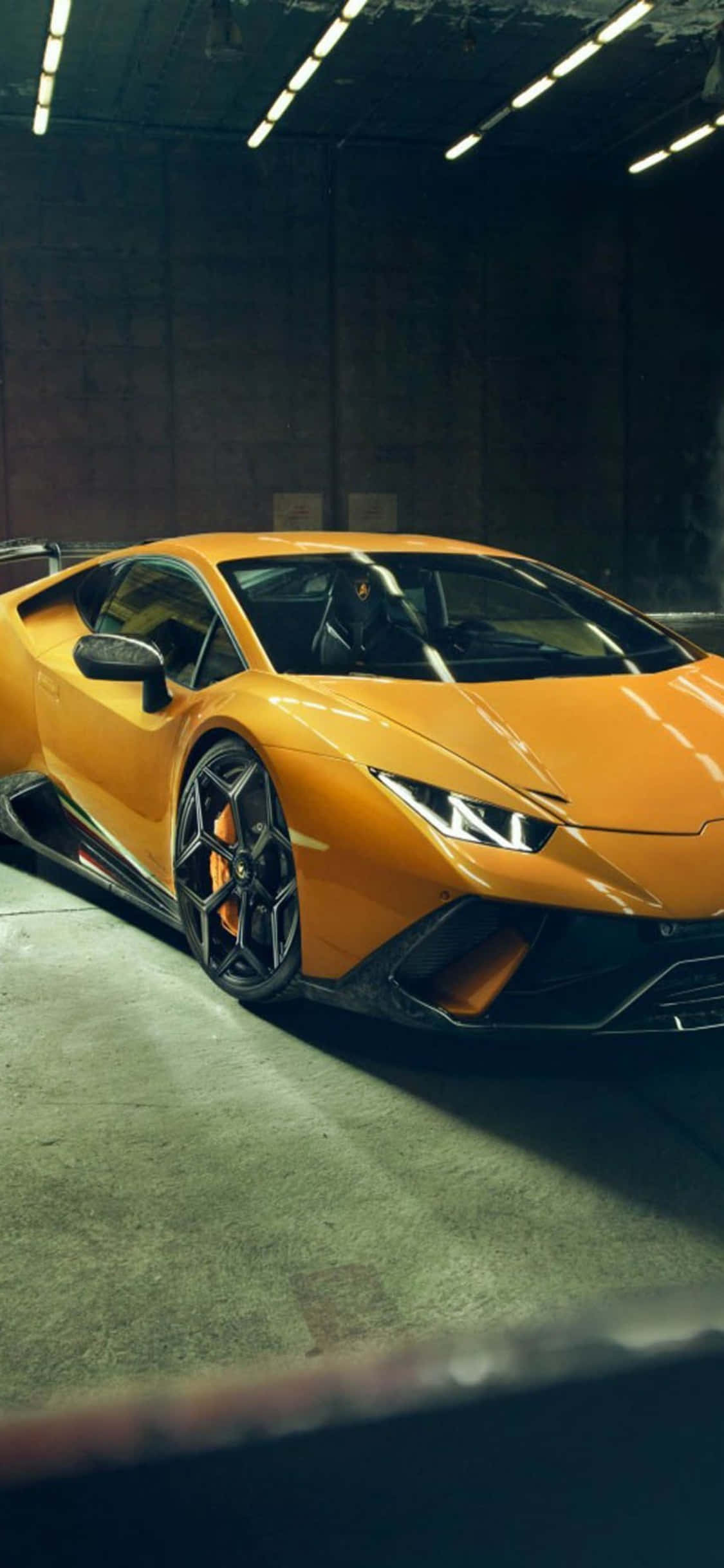 iPhone X Lamborghini 桌布 壁紙 — 一輛鮮橙色的藍寶堅尼 Huracán 停放在燈光昏暗的工業車庫中，天花板的螢光燈投射出銳利的陰影。