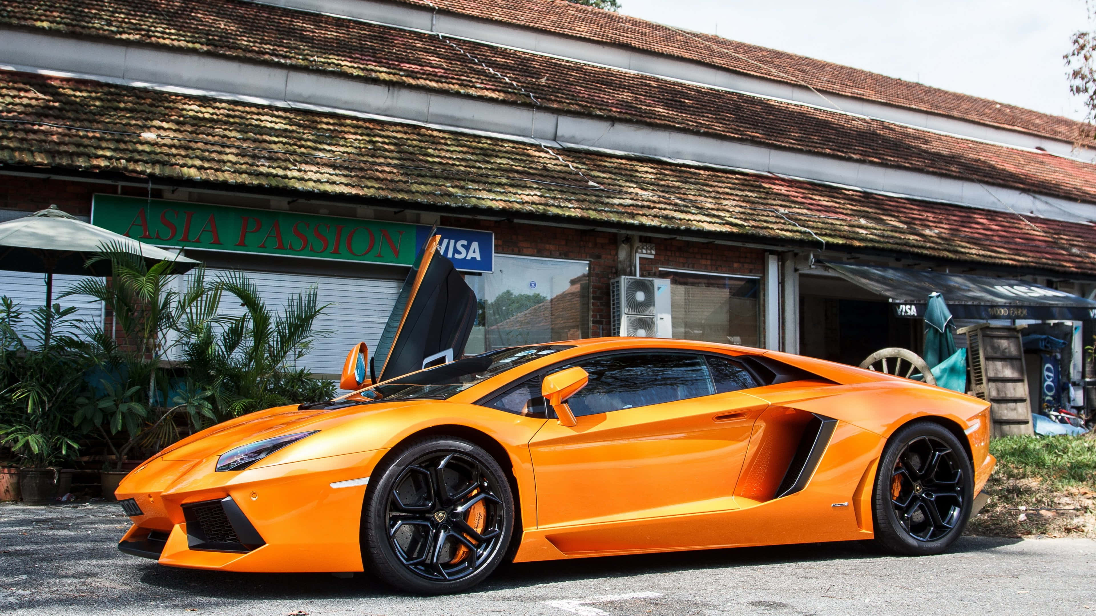 Live Car 桌布 壁紙 — 一輛鮮橙色的藍寶堅尼Aventador，剪刀門打開，停在質樸的磚砌建築前的柏油路上。