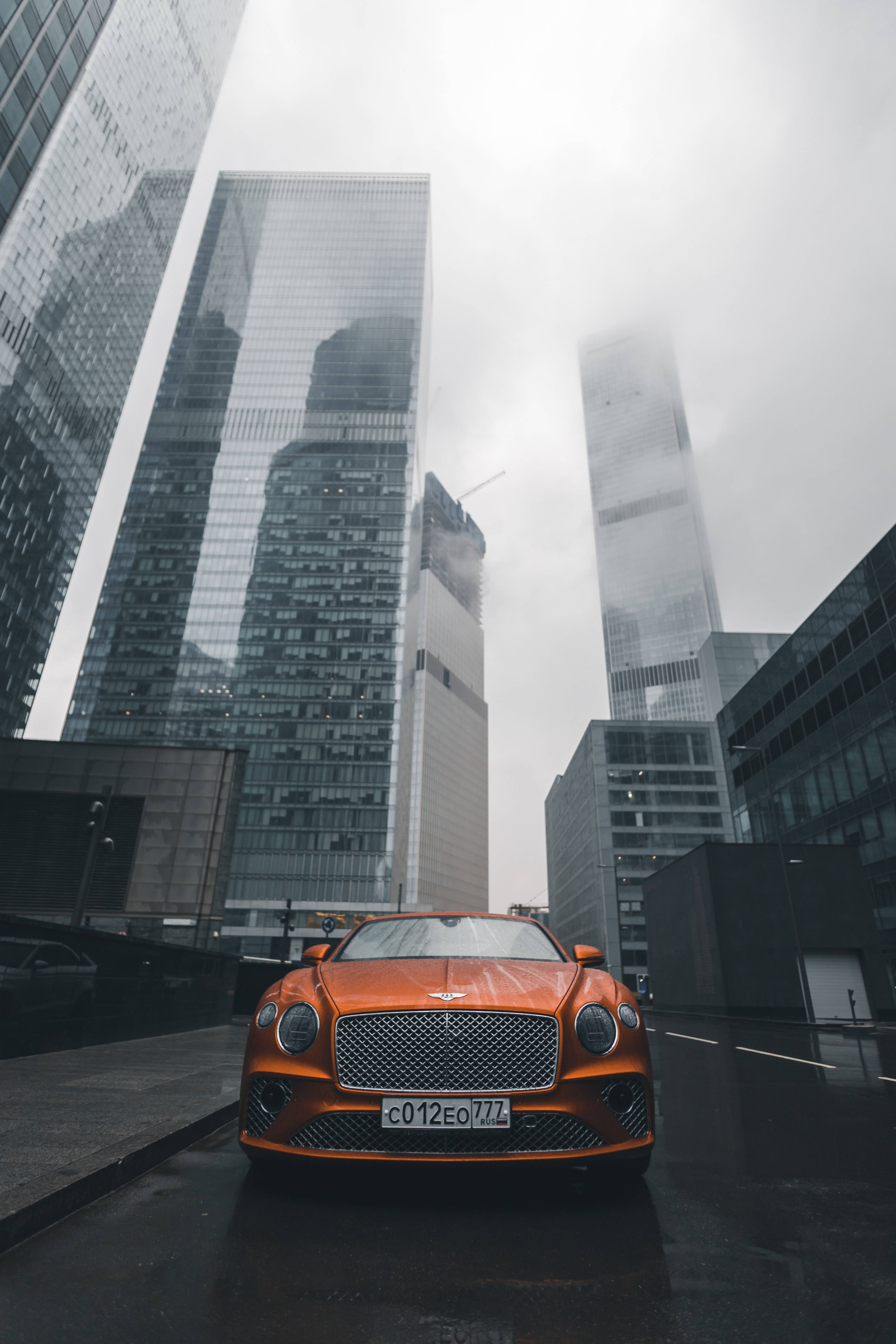 Orange City 桌布 壁紙 — 一輛鮮橙色的賓利 Bentayga 停泊在濕漉漉的都市街道上，周圍是高聳的玻璃摩天大樓，籠罩在迷濛的灰色雲霧中。