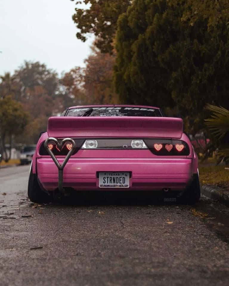 race car 桌布 壁紙 — 一輛鮮粉紅色的改裝車，帶有心形尾燈和心形拖車鉤，停在綠樹成蔭的街道上。