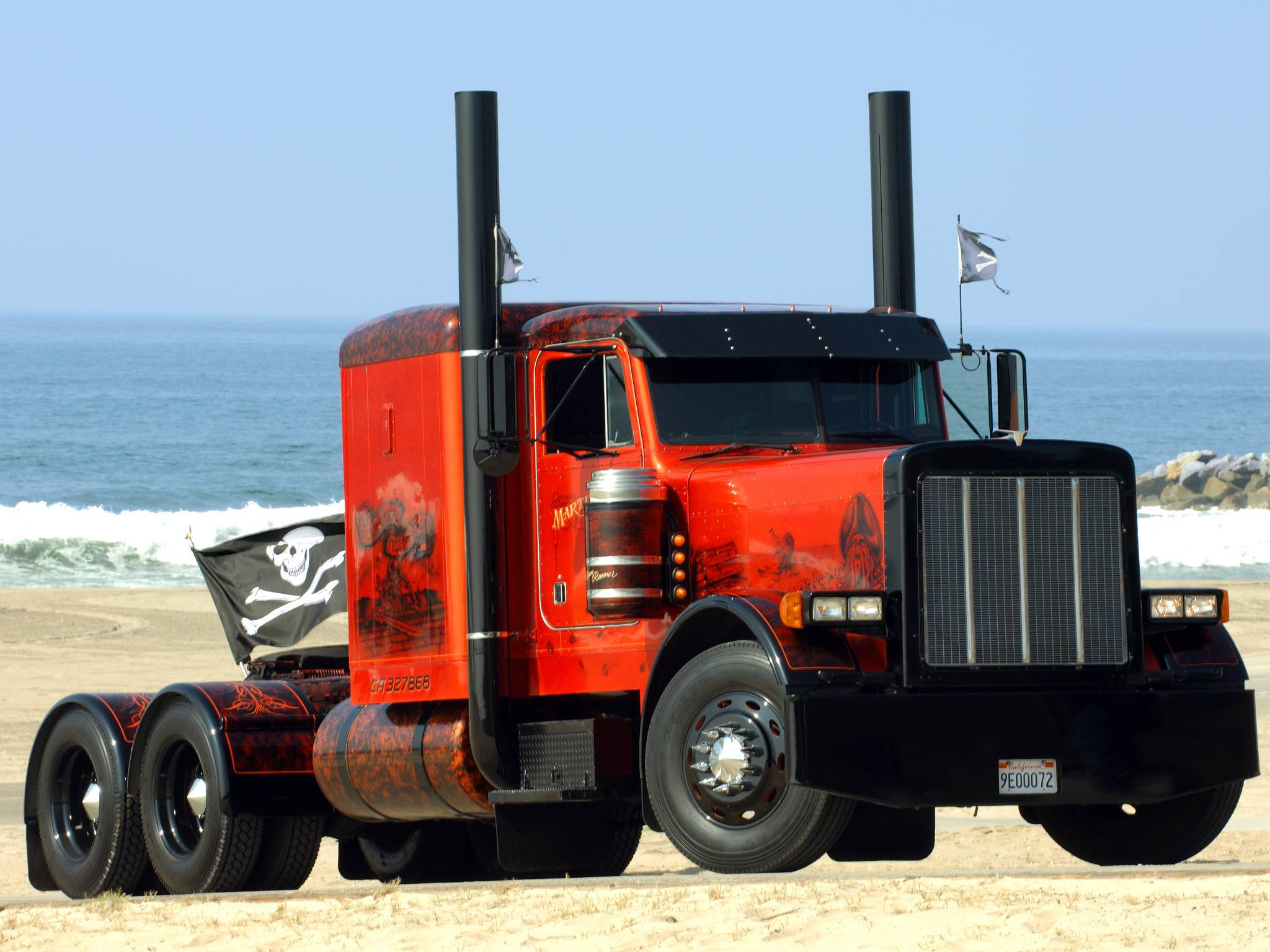 Trucks 桌布 壁紙 — 一輛鮮紅色、帶有海盜主題貼紙和旗幟的定制半掛卡車停泊在沙灘上，旁邊是拍打的海浪。