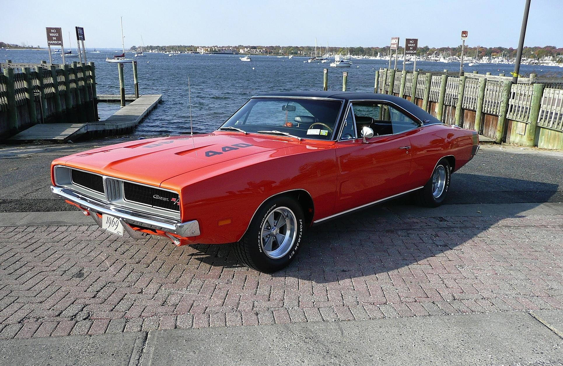 Charger 桌布 壁紙 — 一輛鮮紅色1969年Dodge Charger R/T，搭配黑色乙烯基車頂，停放在磚砌碼頭上，旁邊是停滿船隻的港口，天空清澈湛藍。
