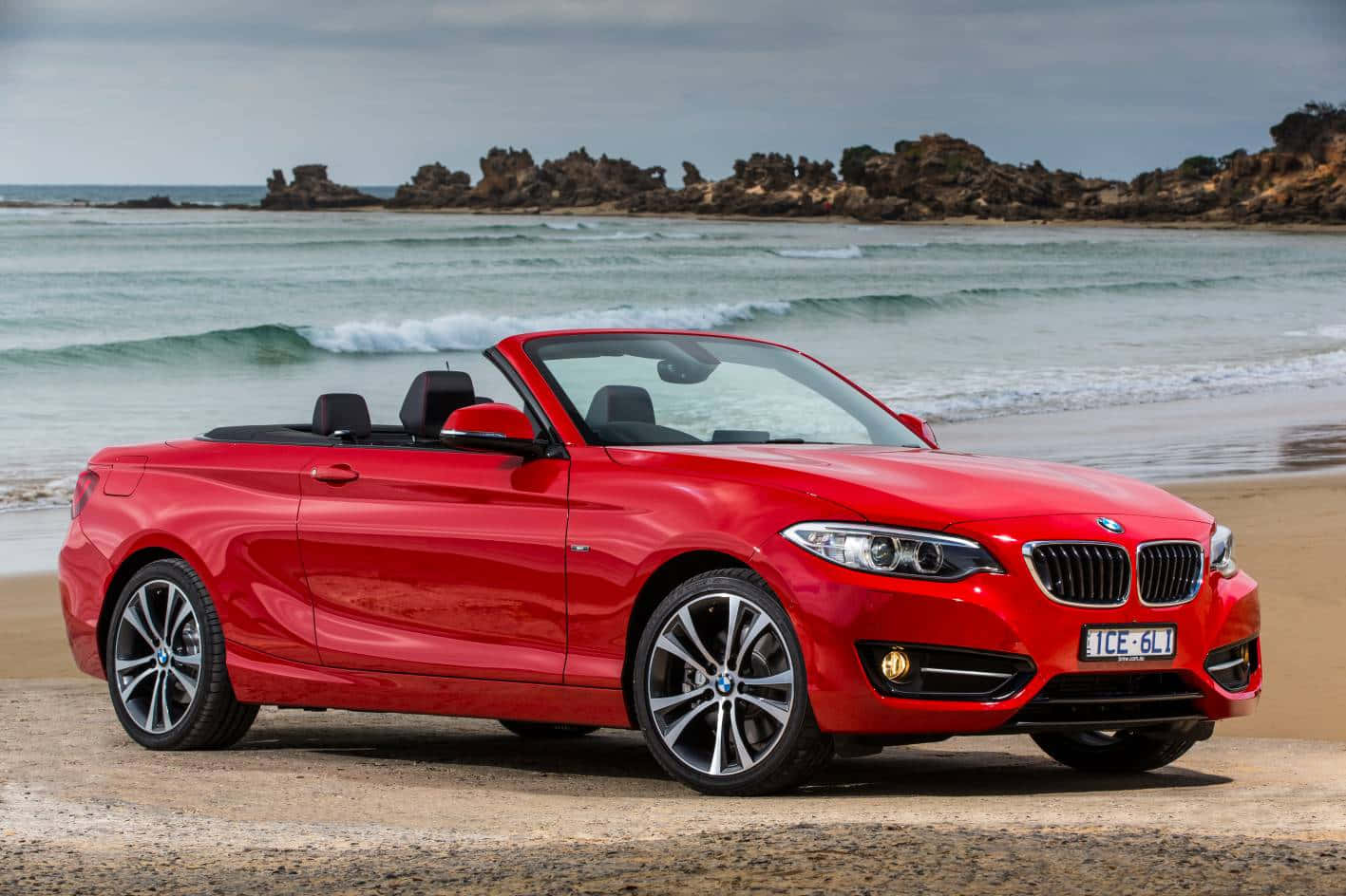 Bmw 2 Series 桌布 壁紙 — 一輛鮮紅色BMW敞篷車停放在沙灘上，背景有海浪和岩石海岬。