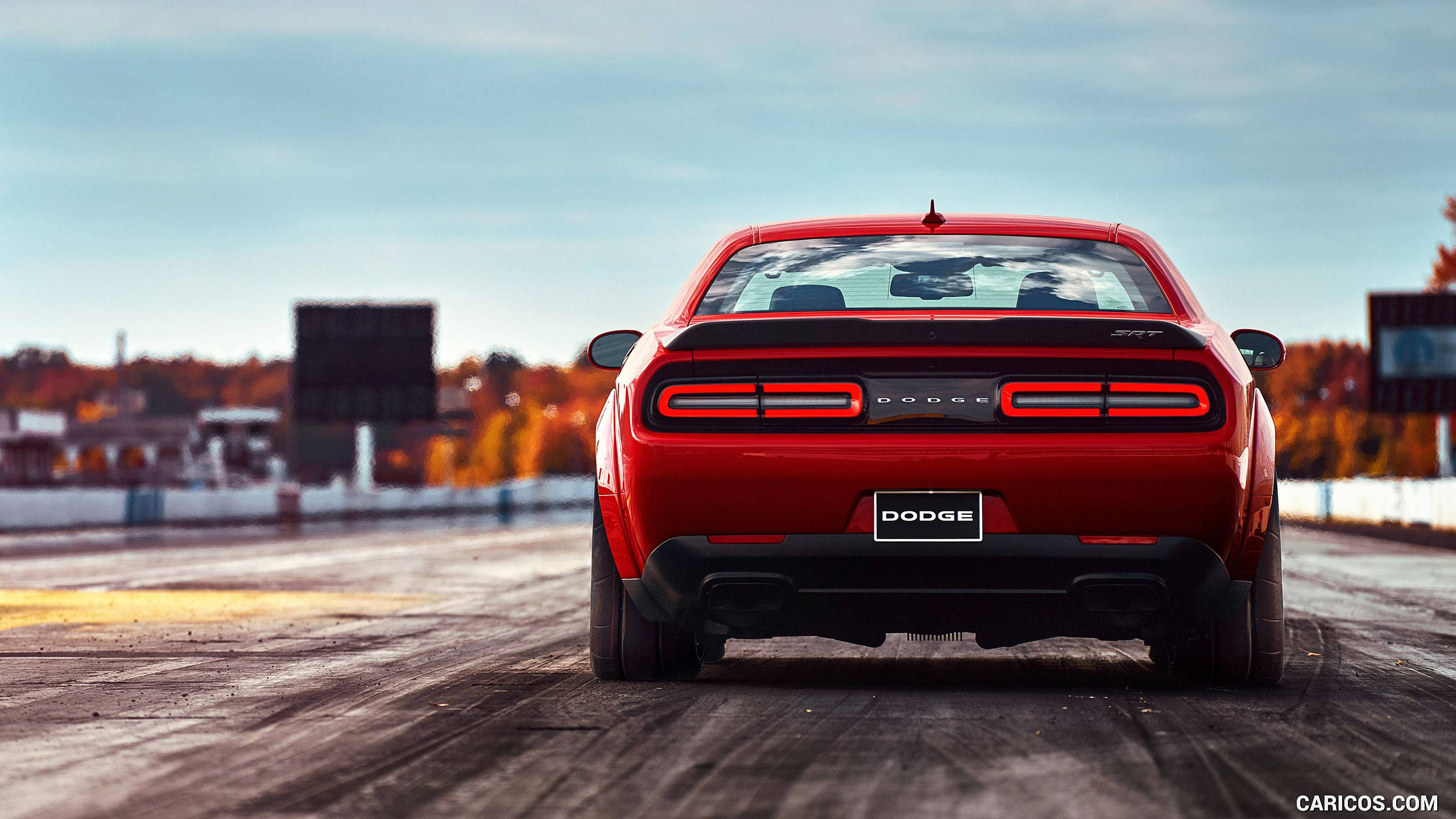 Challenger 桌布 壁紙 — 一輛鮮紅色Dodge Challenger SRT Hellcat在賽道上，背景有秋樹和晴朗天空的後視圖。