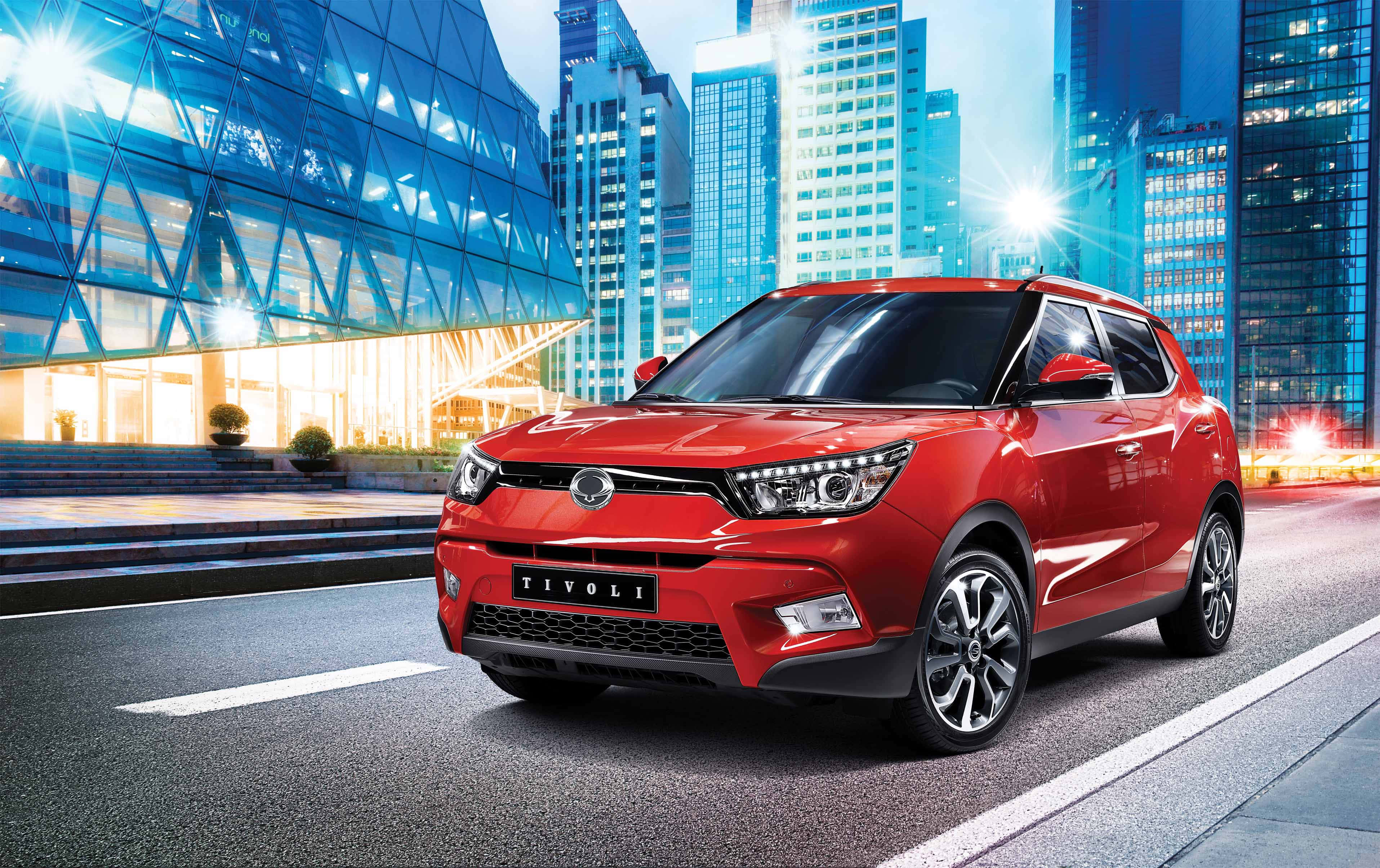 Ssangyong 桌布 壁紙 — 一輛鮮紅色SUV停在閃亮的城市街道上，高聳的玻璃摩天大樓在暮色燈光下閃耀。