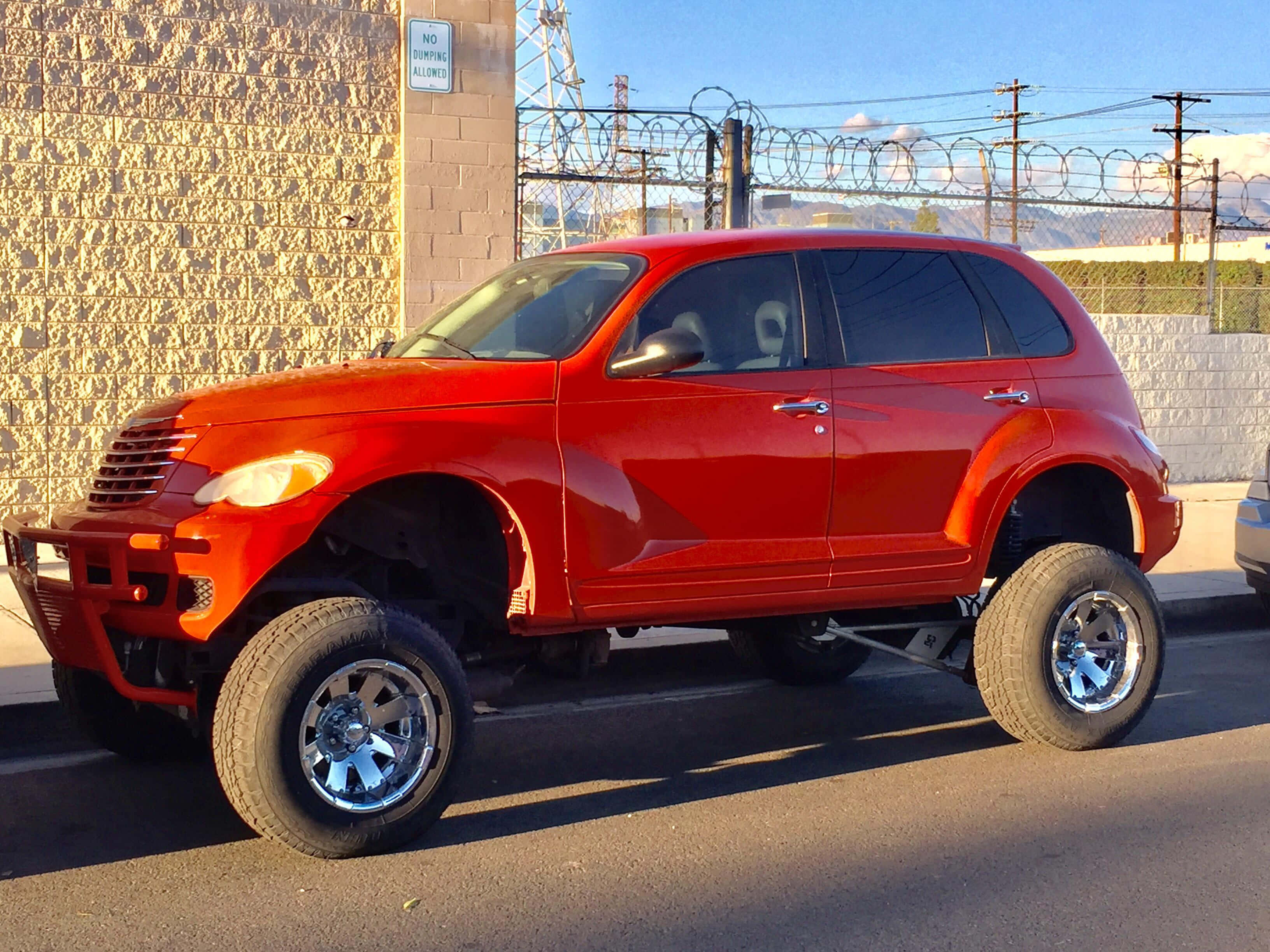 Chrysler Pt Cruiser 桌布 壁紙 — 一輛鮮紅色升高底盤的Chrysler PT Cruiser，配備大型鍍鉻輪圈，停泊在陽光明媚的街道上，旁邊是石牆和鐵絲網圍欄。