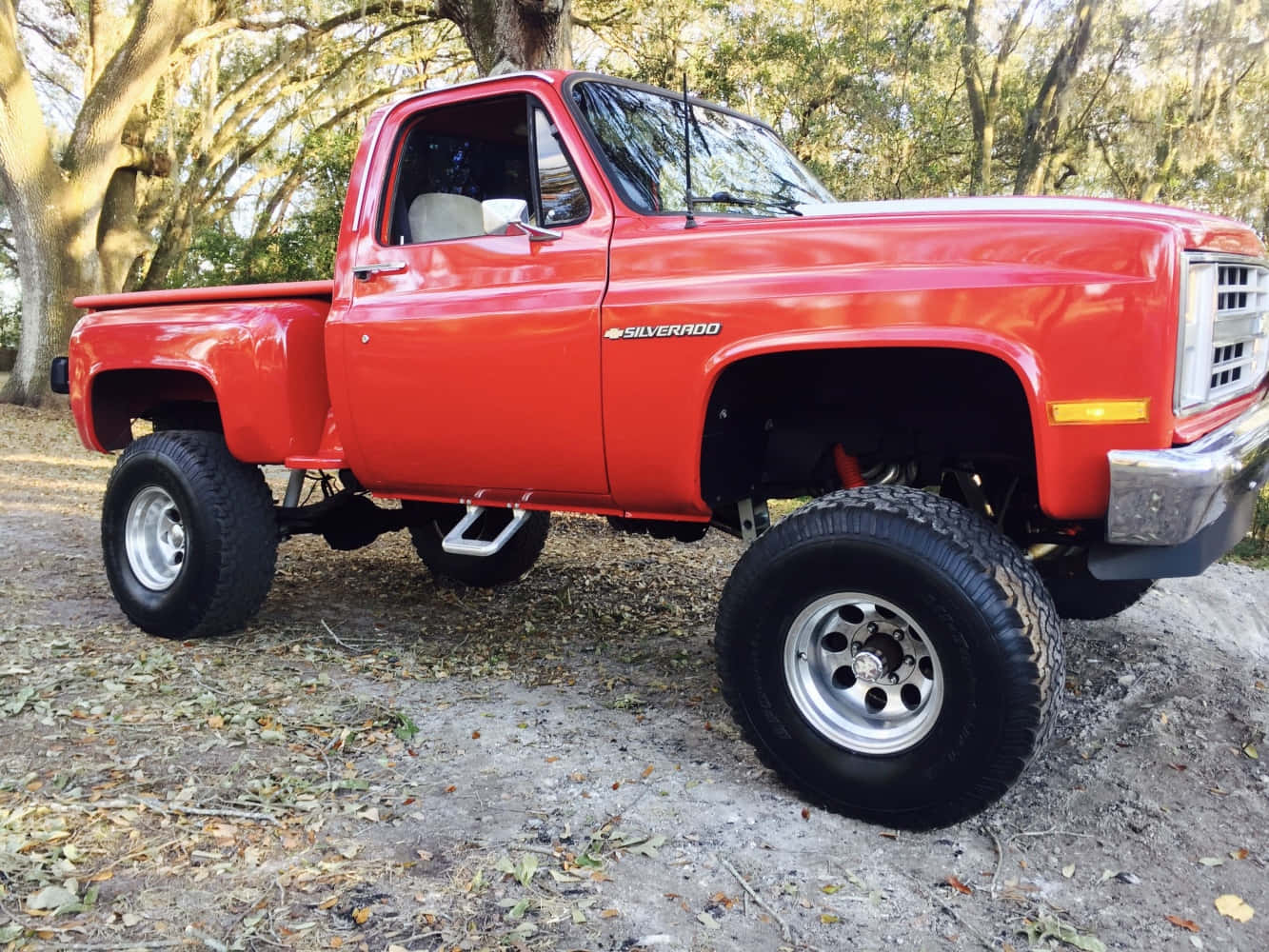 Square Body Truck 桌布 壁紙 — 一輛鮮紅色升高底盤的雪佛蘭Silverado皮卡車，配備大型輪胎，停在高大樹木下鋪滿落葉的泥路上。
