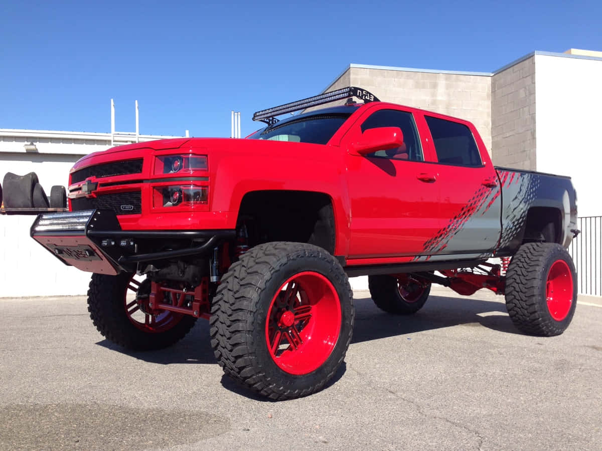 Jacked Up Trucks 桌布 壁紙 — 一輛鮮紅色升高底盤的雪佛蘭皮卡車，配備超大輪胎和定制圖案，停在晴朗藍天下的瀝青路面上。
