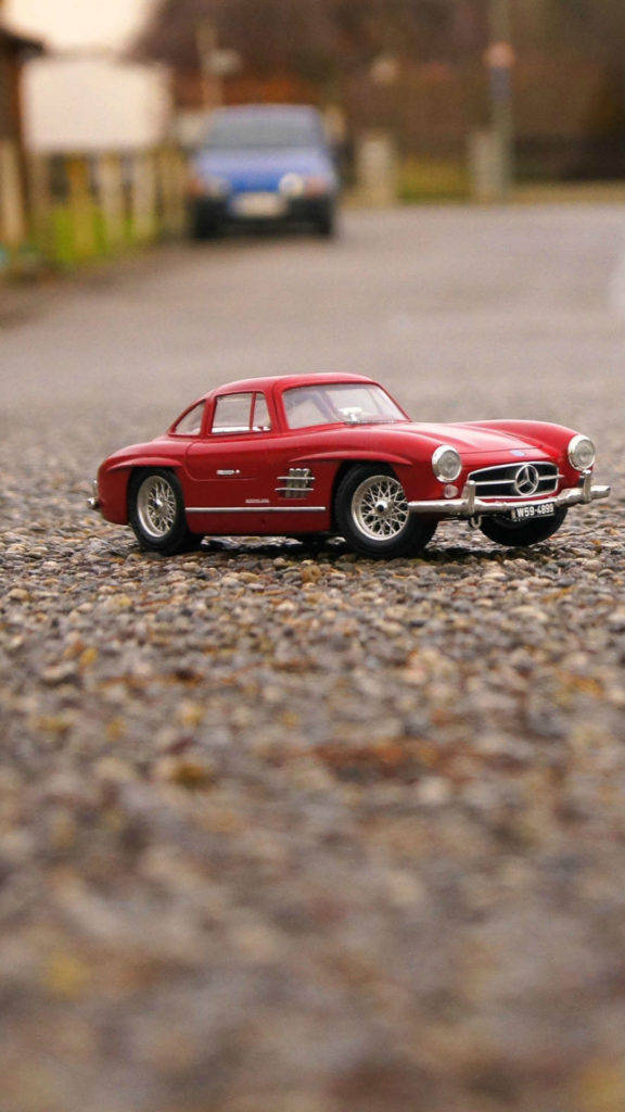 汽車 iPhone 壁紙 桌布 壁紙 — 一輛鮮紅色復古Mercedes-Benz玩具車停在碎石路上，背景有一輛模糊的藍色汽車。