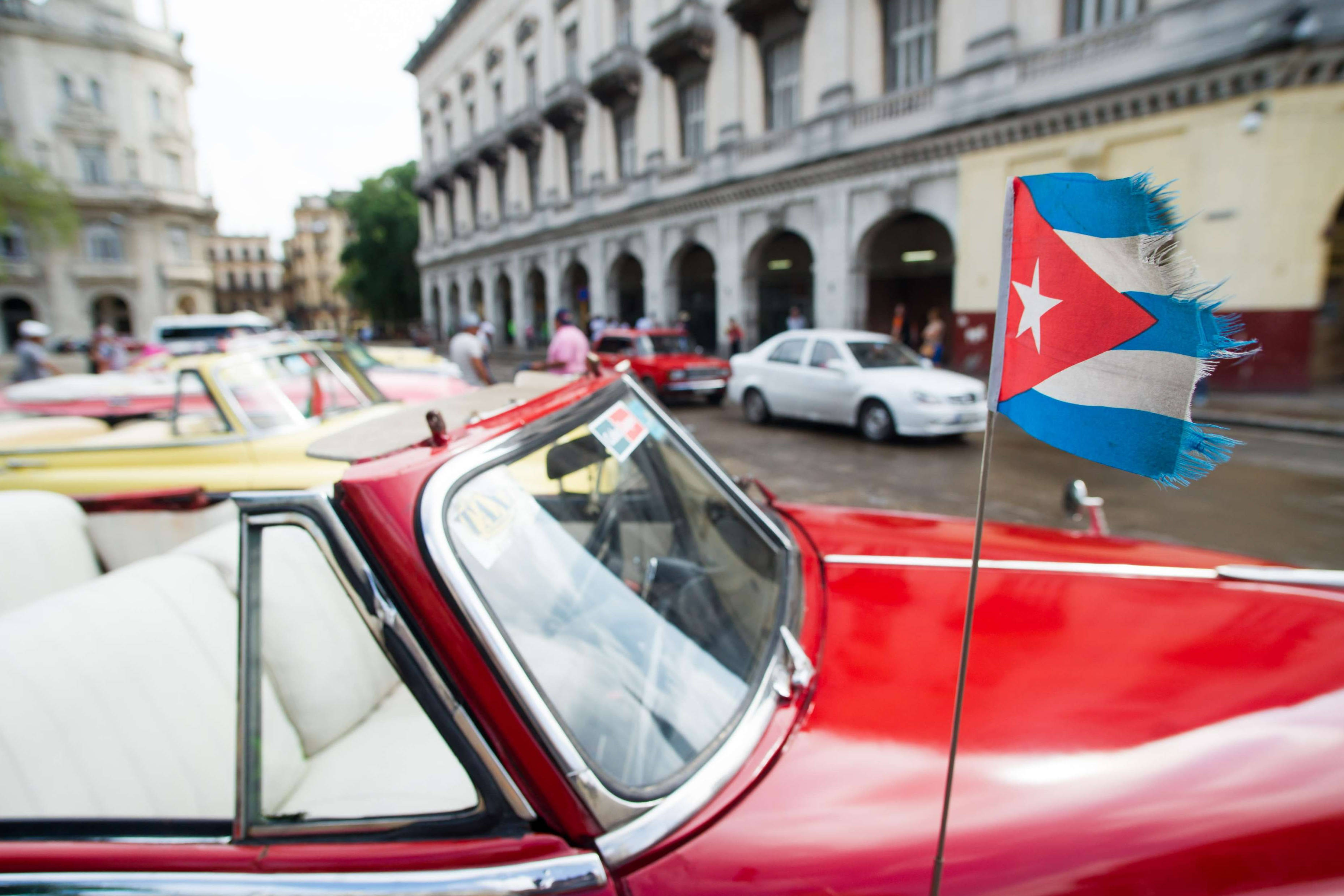 Cuban 桌布 壁紙 — 一輛鮮紅色復古敞篷車，引擎蓋上掛著破舊的古巴國旗，停放在繁忙的殖民時期街道上。