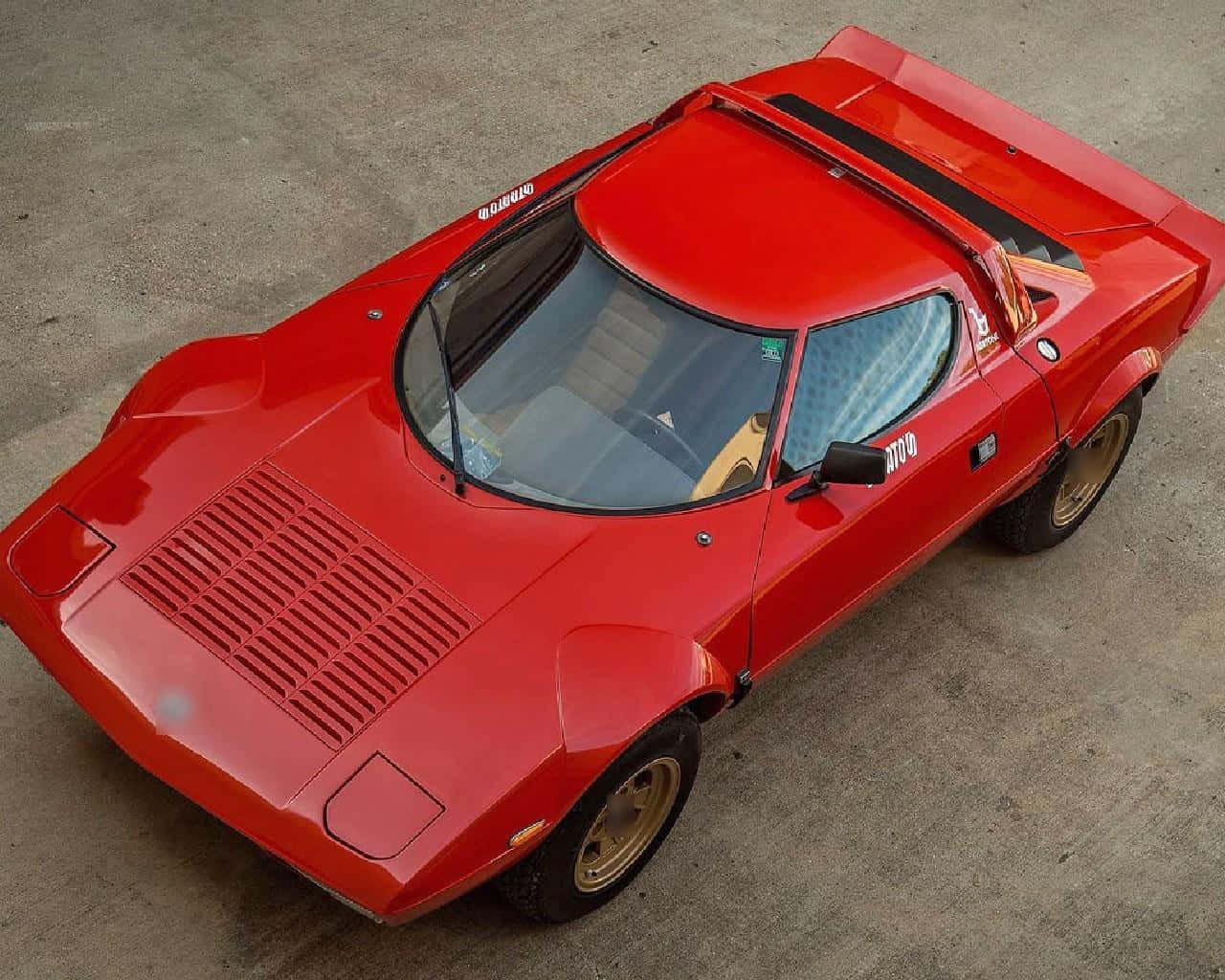 Lancia 桌布 壁紙 — 一輛鮮紅色復古跑車配金色輪胎，停在混凝土路面上，在明亮的日光下。
