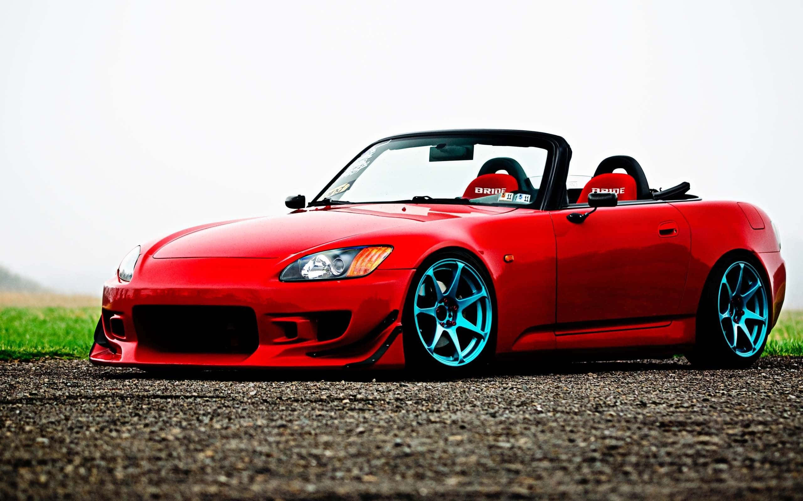 Honda S2000 桌布 壁紙 — 一輛鮮紅色敞篷跑車，搭配搶眼的藍色輪圈，停放在碎石地上，背景是朦朧的綠色田野。
