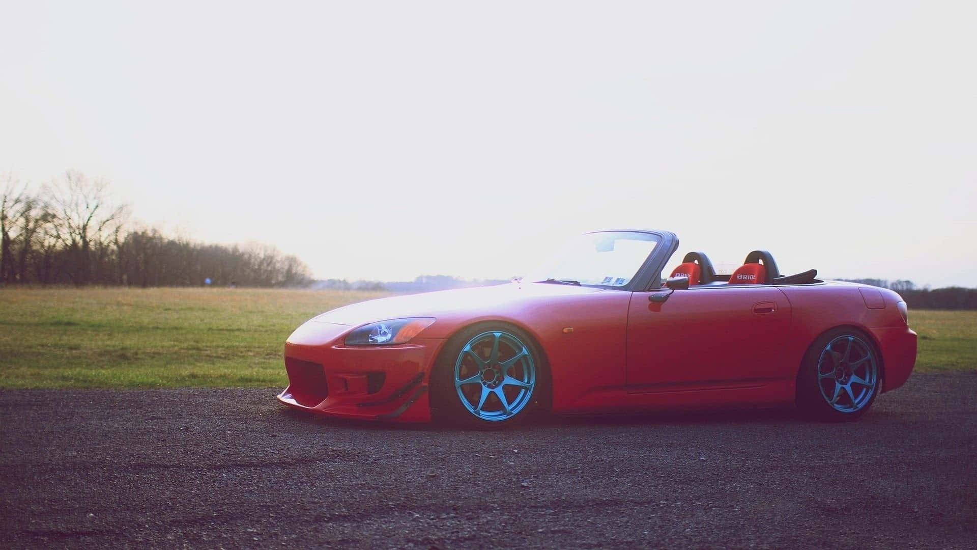 Honda S2000 桌布 壁紙 — 一輛鮮紅色敞篷跑車搭配藍色輪圈，停放在日落時分草地上的碎石路上。