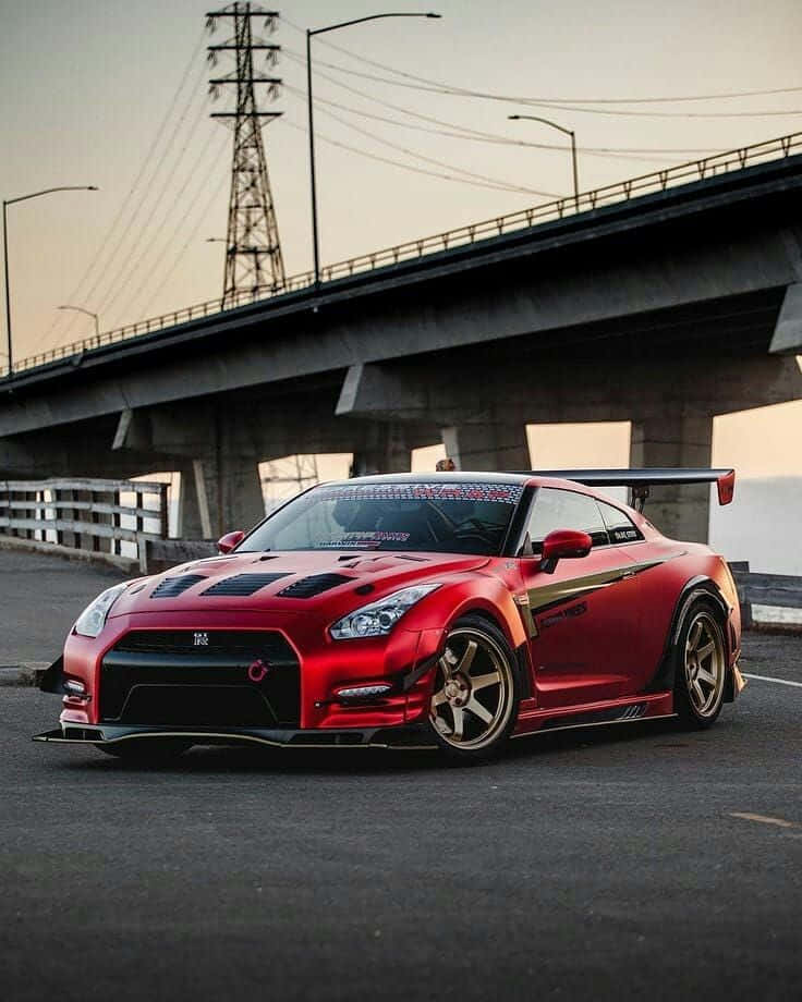 Nissan 桌布 壁紙 — 一輛鮮紅色日產GT-R跑車，帶金色點綴，在黃昏時停泊在混凝土天橋下。