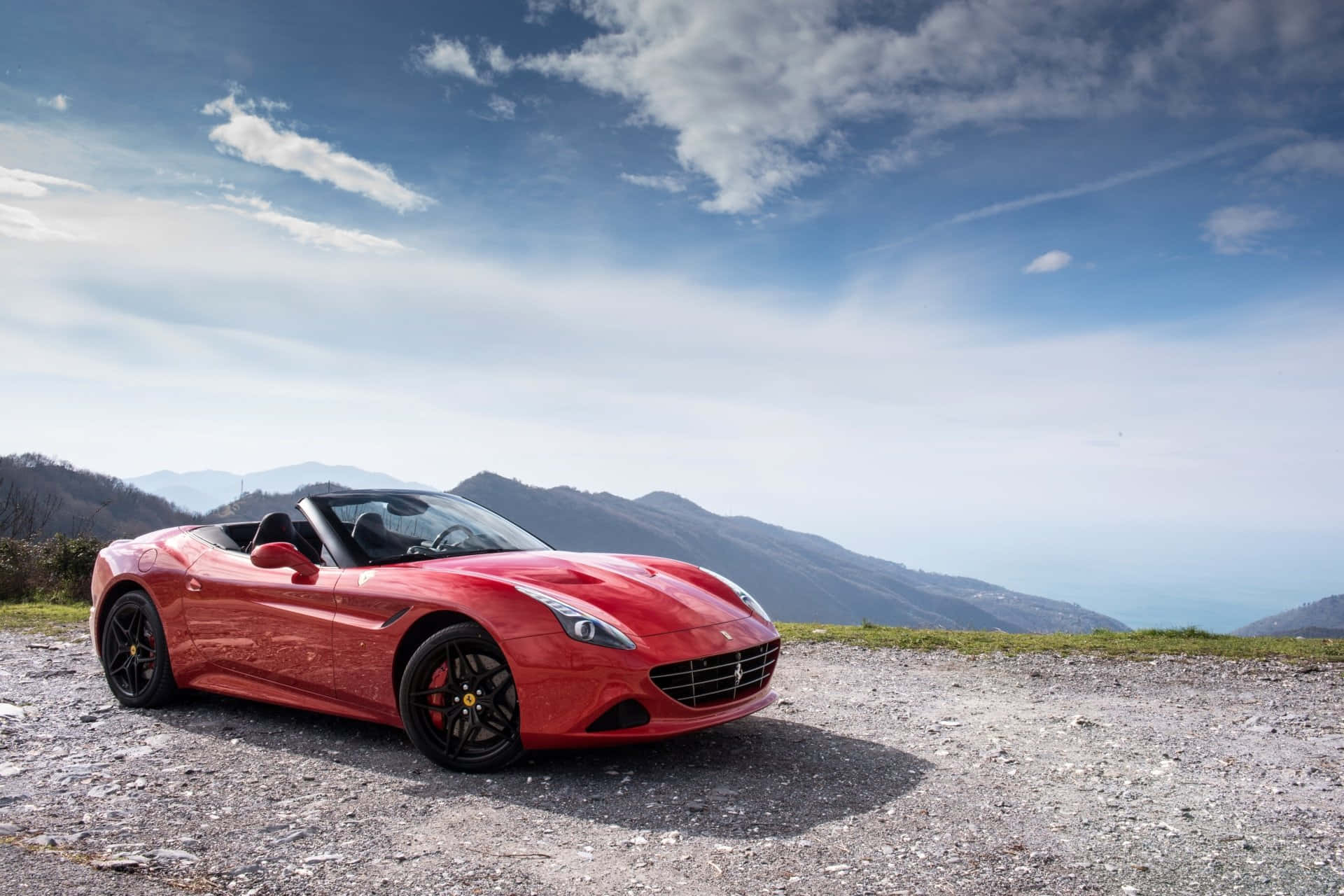 Ferrari California T 桌布 壁紙 — 一輛鮮紅色法拉利敞篷車停泊在碎石觀景點，背景是連綿起伏的山巒和點綴著白雲的藍天。