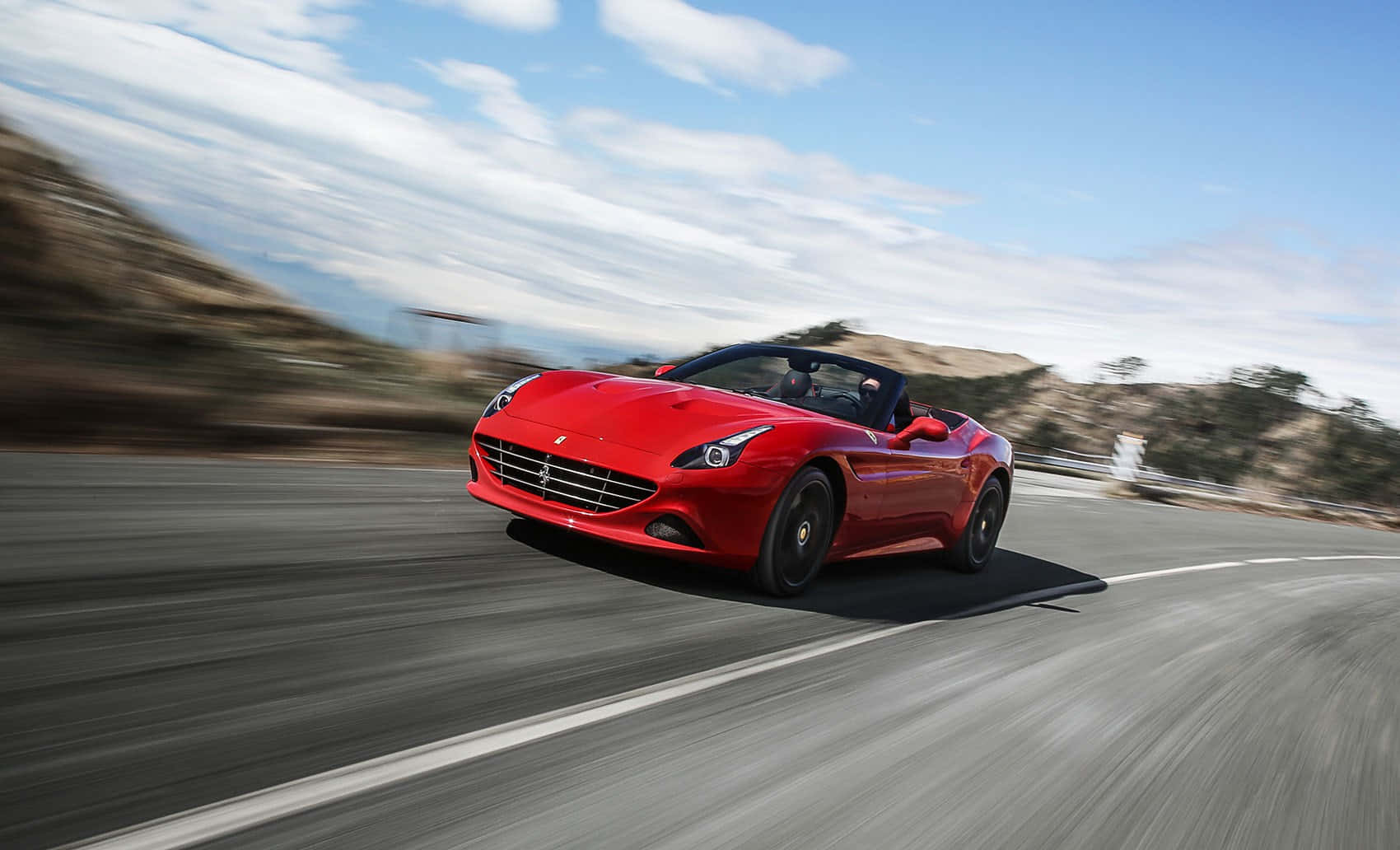 Ferrari California T 桌布 壁紙 — 一輛鮮紅色法拉利敞篷車在晴朗藍天白雲下，沿著蜿蜒的山路飛馳。