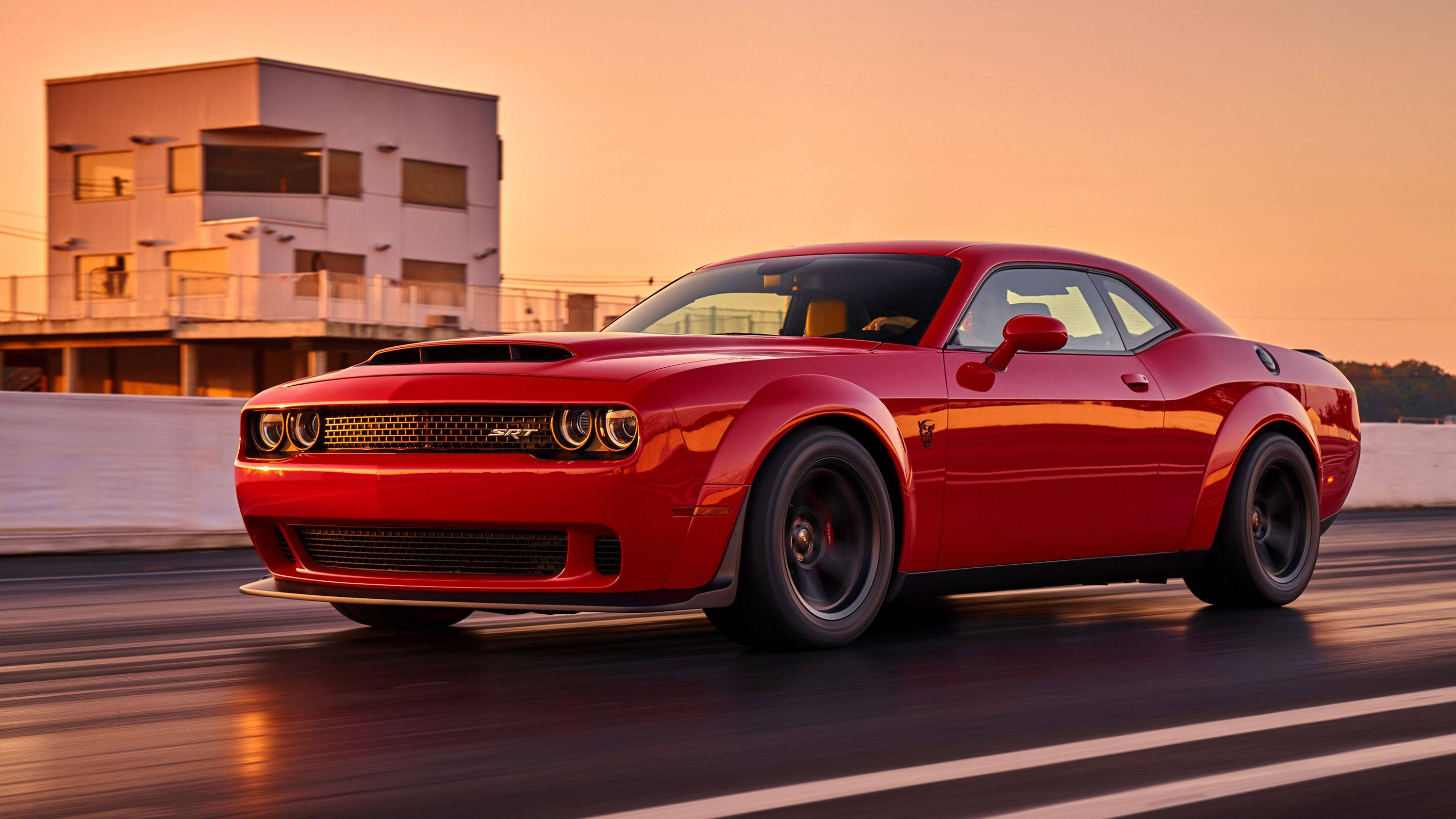 Dodge 桌布 壁紙 — 一輛鮮紅色的 Dodge Challenger 在日落時分沿著賽道飛馳，背景是一座現代建築。