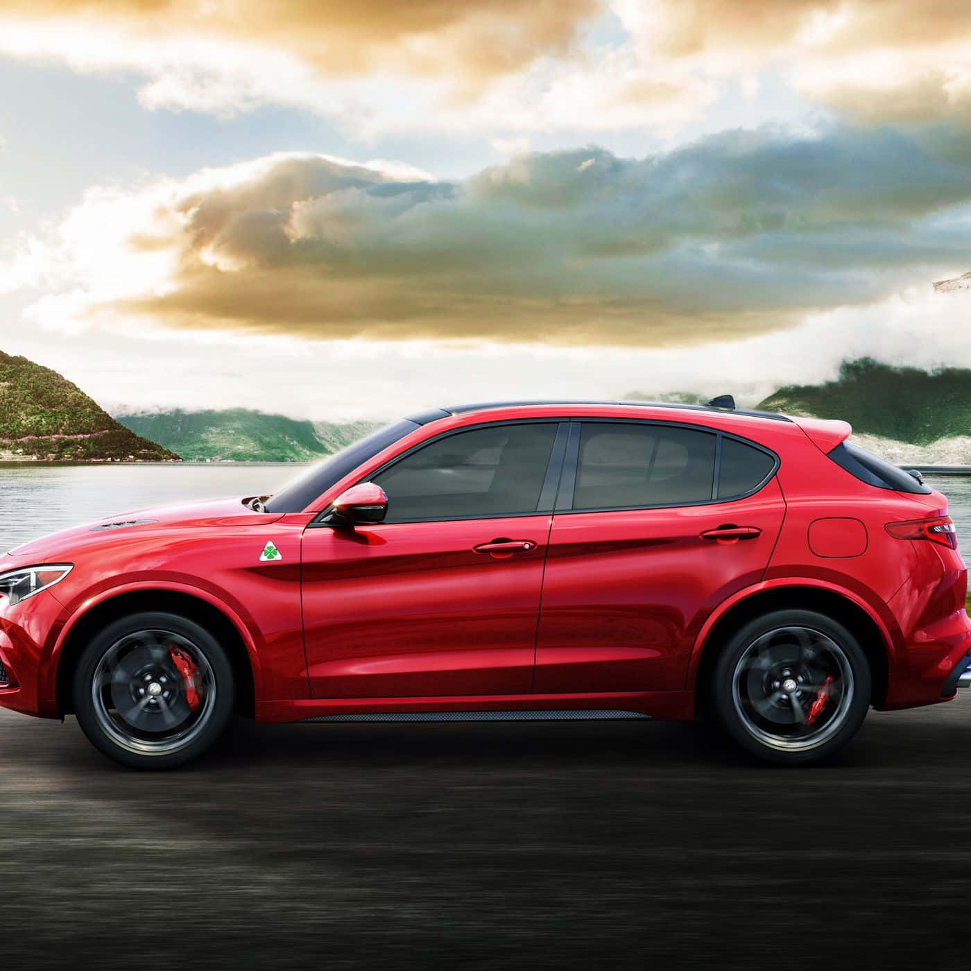 Alfa Romeo Stelvio 桌布 壁紙 — 一輛鮮紅色的Alfa Romeo Stelvio SUV沿著湖邊道路行駛，背景是霧氣繚繞的山脈和金色的雲彩。