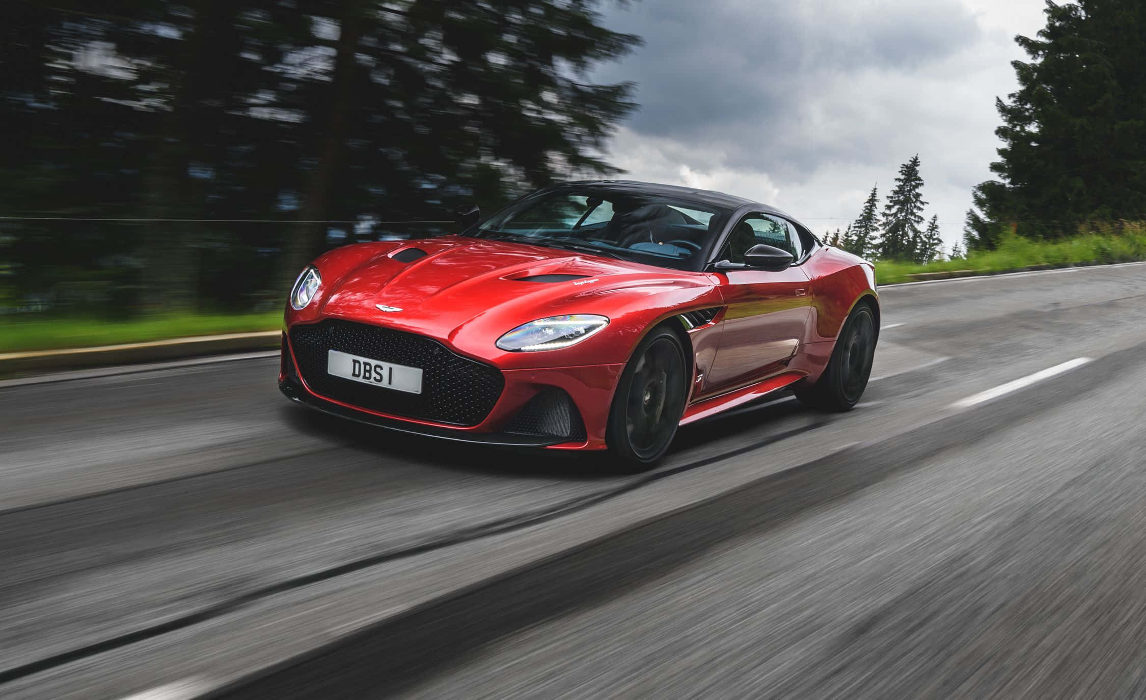 奧斯頓馬丁 桌布 壁紙 — 一輛鮮紅色的Aston Martin DBS1超級跑車沿著蜿蜒的林間道路疾馳，動態模糊強調了其速度。