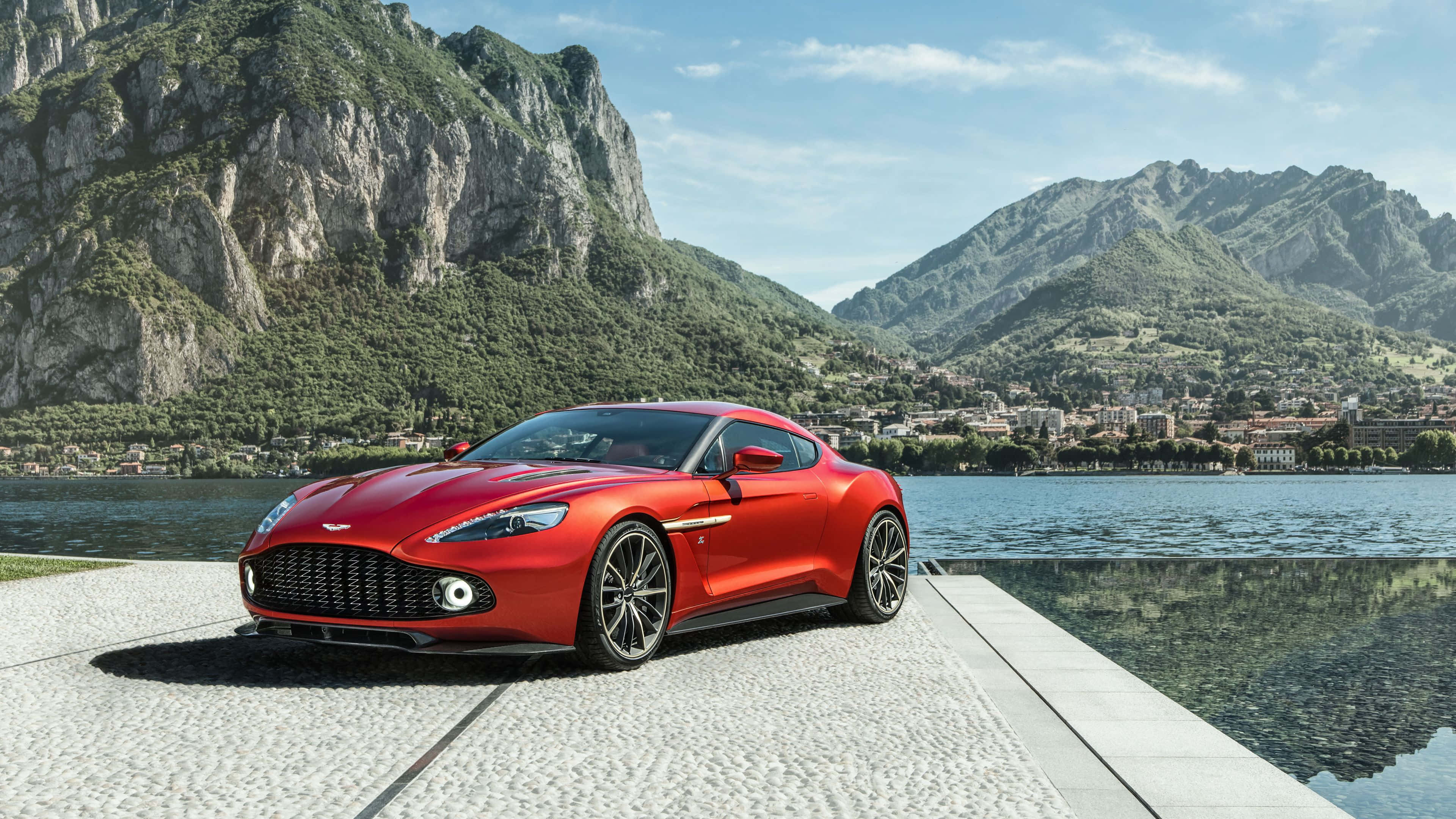 奧斯頓馬丁 桌布 壁紙 — 一輛鮮紅色的Aston Martin跑車在湖邊碼頭上閃閃發光，背景是崎嶇的山脈和一個小鎮，在清澈的藍天下。