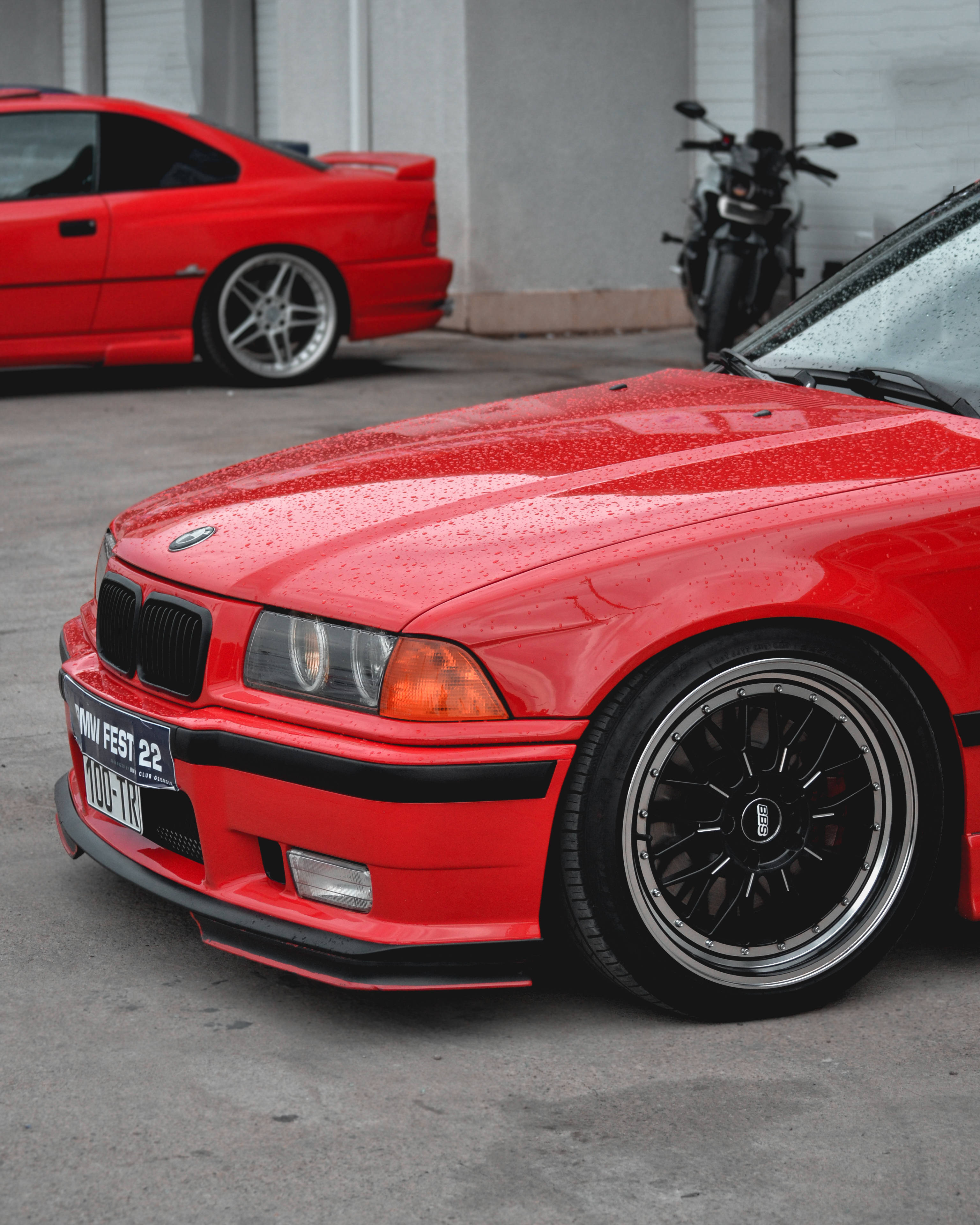 Racing 桌布 壁紙 — 一輛鮮紅色的BMW E36雙門轎跑車，在柔和光線下雨滴閃閃發光，停在混凝土車庫中一輛摩托車旁邊。