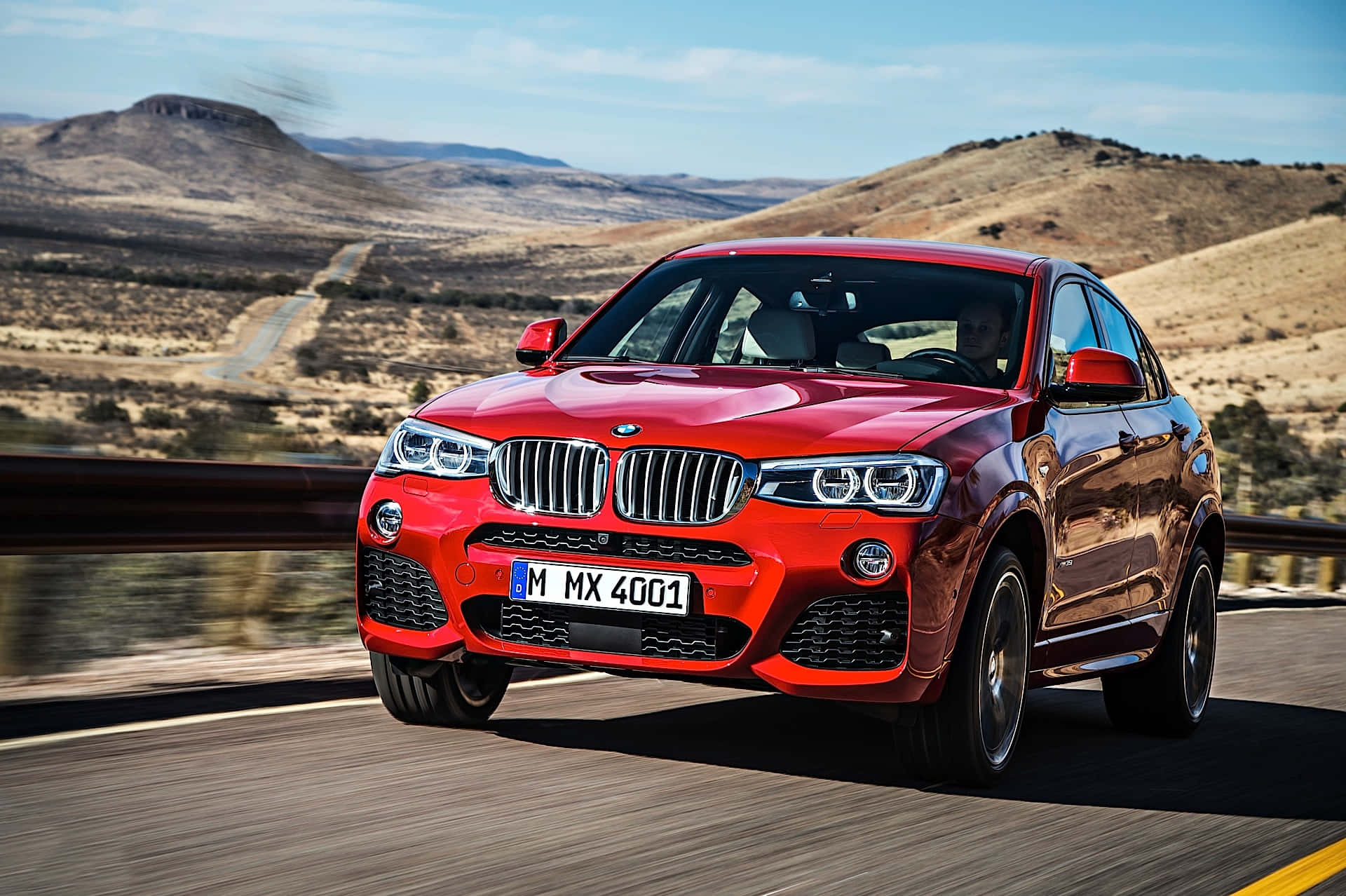 Bmw X4 桌布 壁紙 — 一輛鮮紅色的BMW X4 M40i在晴朗藍天下，沿著崎嶇山丘的沙漠高速公路飛馳。