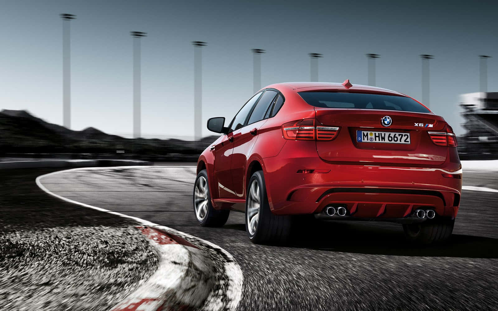 BMW X6 M 桌布 壁紙 — 一輛鮮紅色的BMW X6 M在賽道彎道高速奔馳，瀝青路面呈現動態模糊效果，背景有高聳的燈柱。
