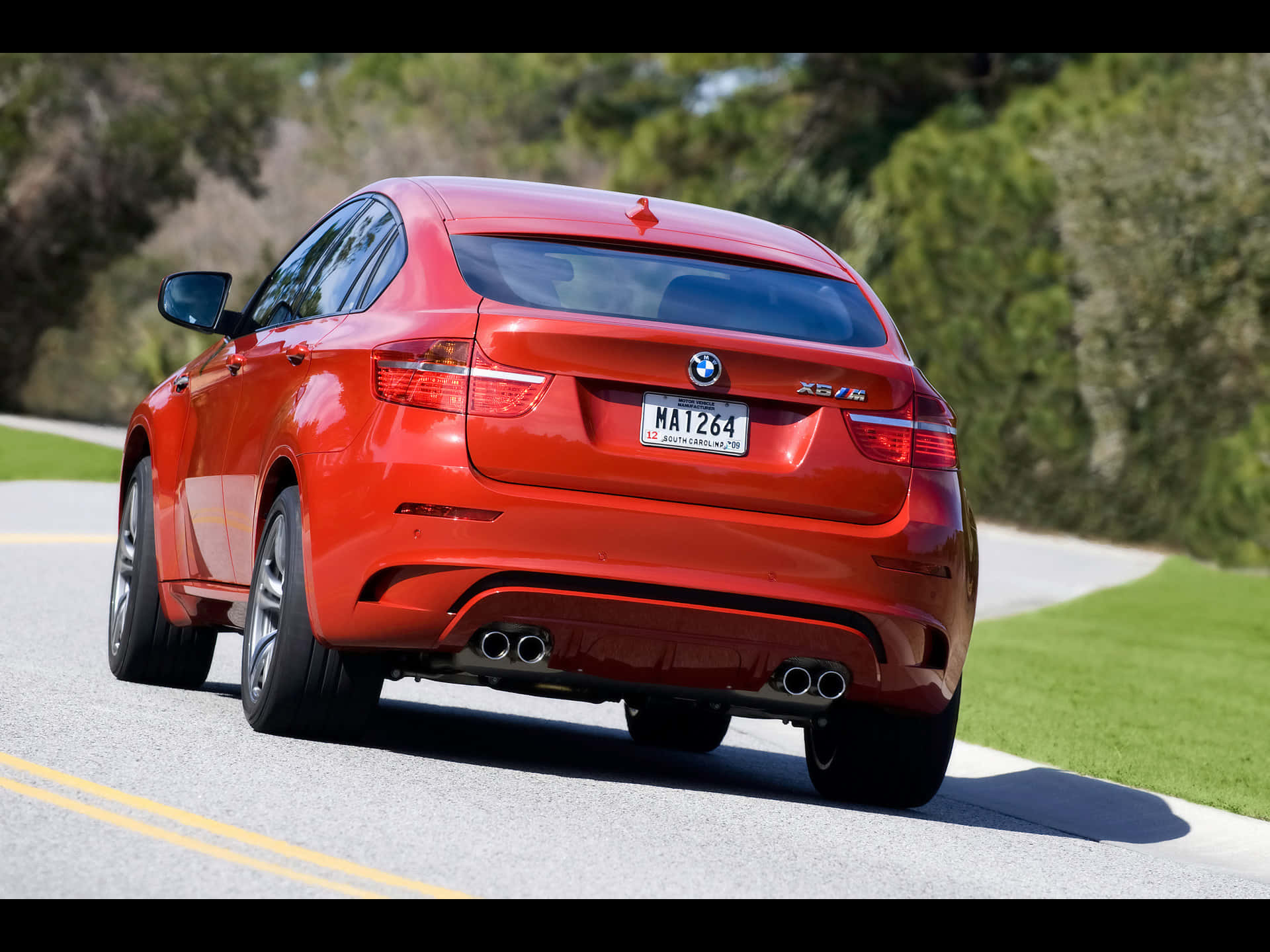 BMW X6 M 桌布 壁紙 — 一輛鮮紅色的BMW X6 SUV沿著陽光明媚的道路行駛，道路兩旁綠樹成蔭，草地茂盛。