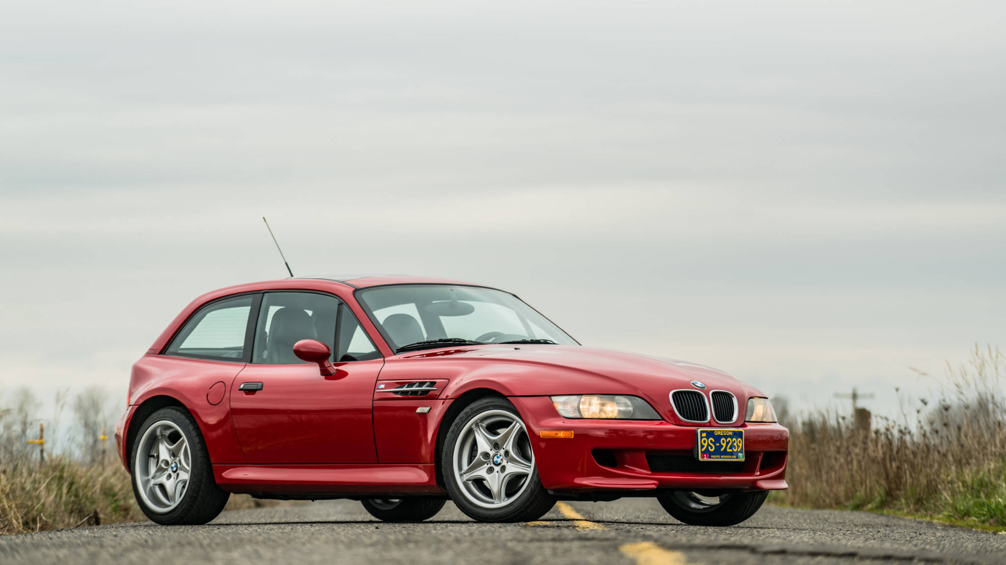 m 桌布 壁紙 — 一輛鮮紅色的BMW Z3敞篷跑車停泊在鄉間的柏油路上，天空多雲，背景有乾草。