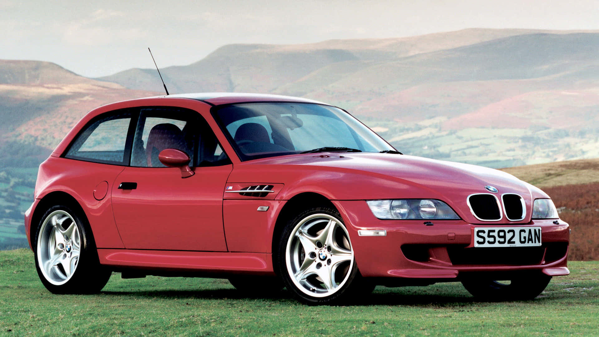 Bmw Z3 桌布 壁紙 — 一輛鮮紅色的BMW Z3跑車停在草地上，背景是起伏的丘陵，天空柔和。