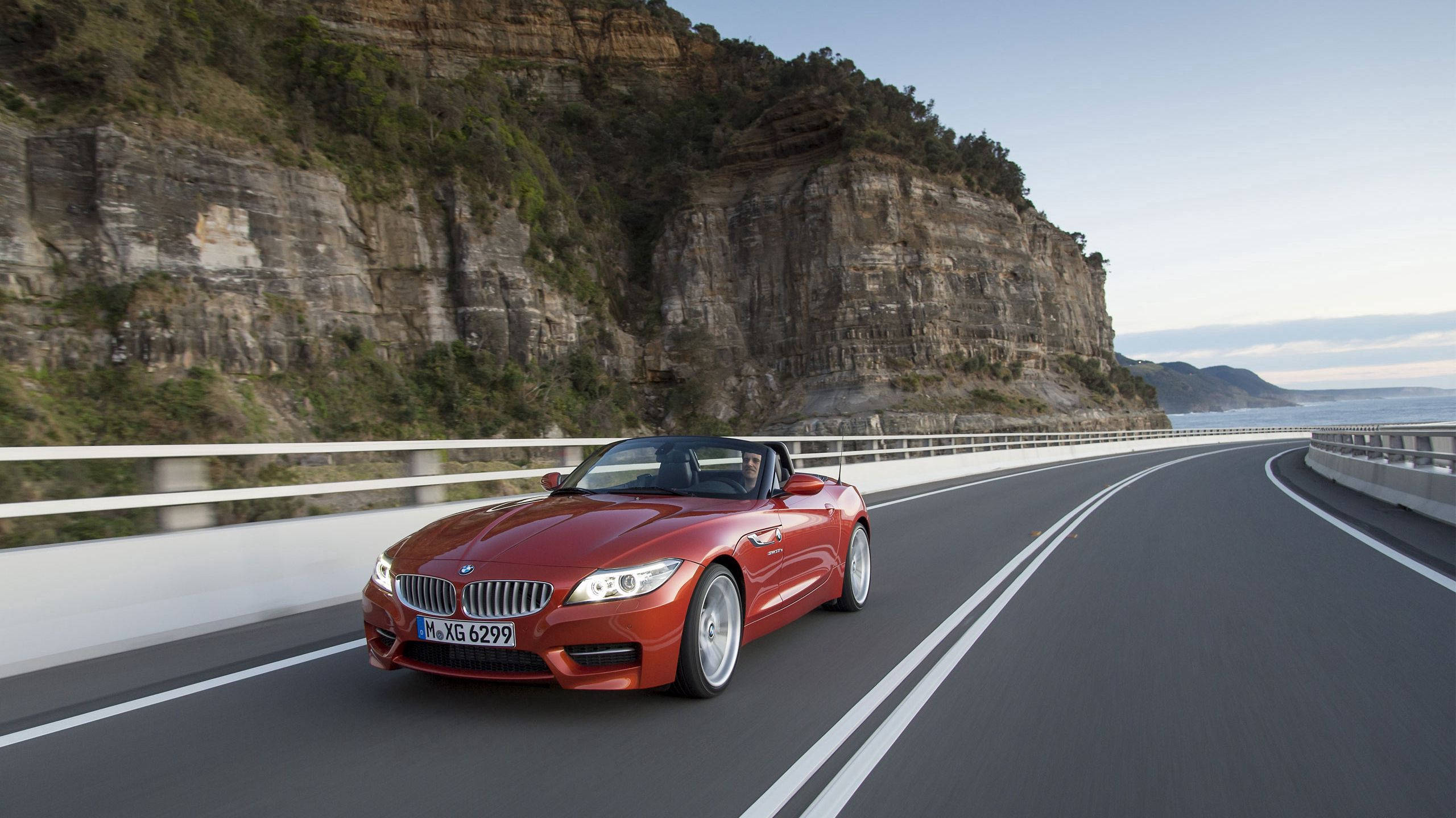 BMW 動圖 桌布 壁紙 — 一輛鮮紅色的BMW Z4敞篷跑車沿著海岸公路行駛，兩旁是崎嶇的懸崖和海洋景觀，天空晴朗。