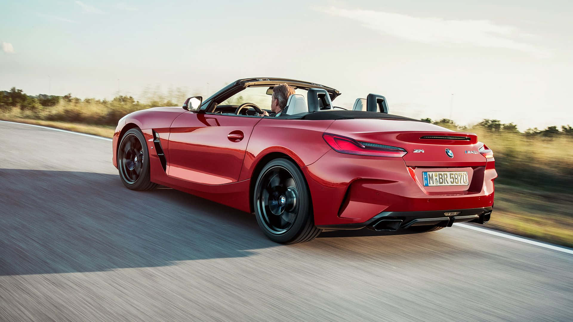 Bmw Z4 桌布 壁紙 — 一輛鮮紅色的BMW Z4敞篷跑車在陽光明媚的道路上行駛，背景有模糊的動態和金色的樹葉。