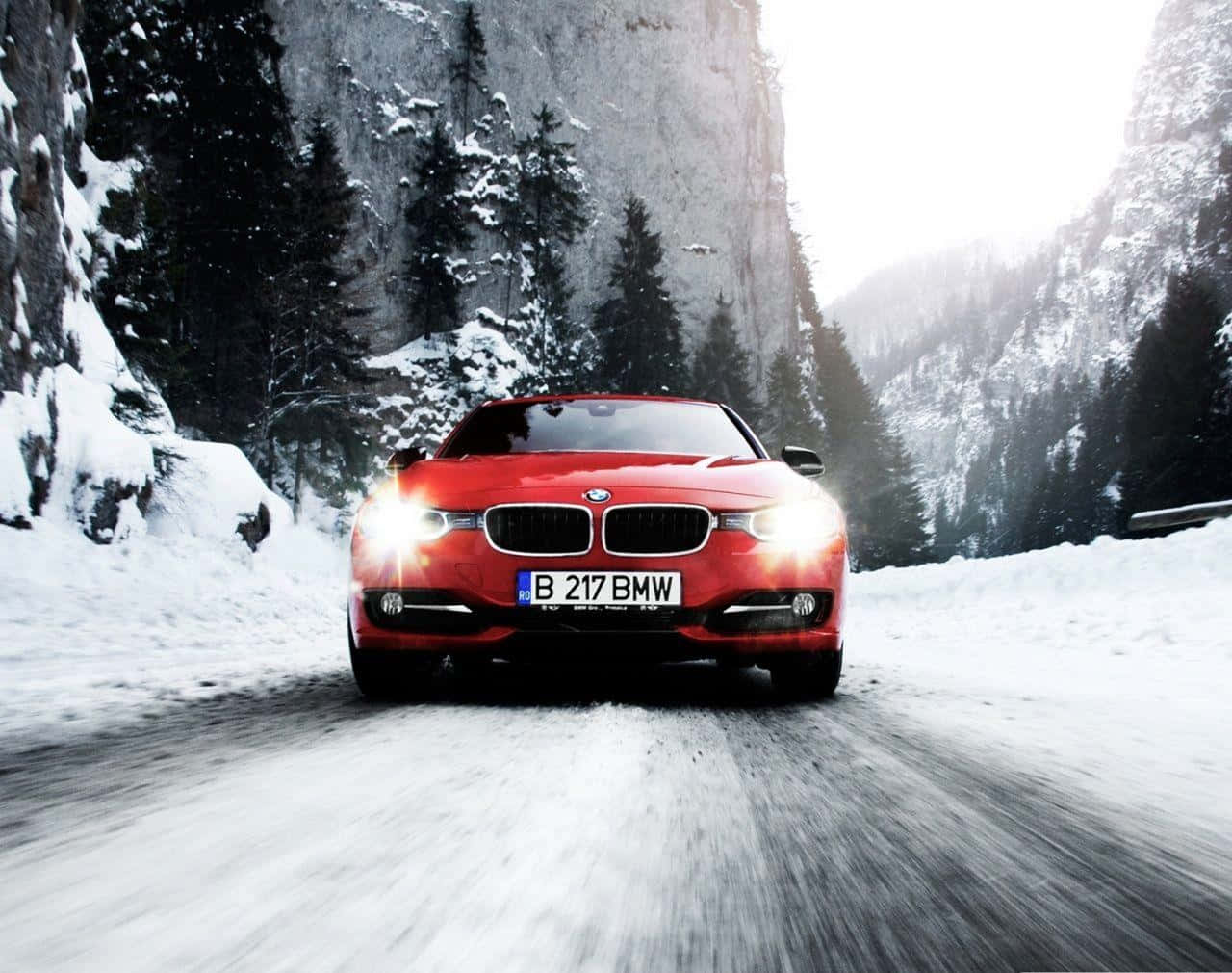 classic bmw 桌布 壁紙 — 一輛鮮紅色的BMW跑車在白雪覆蓋的山路上飛馳，兩旁是高聳的常綠樹木和岩石峭壁。