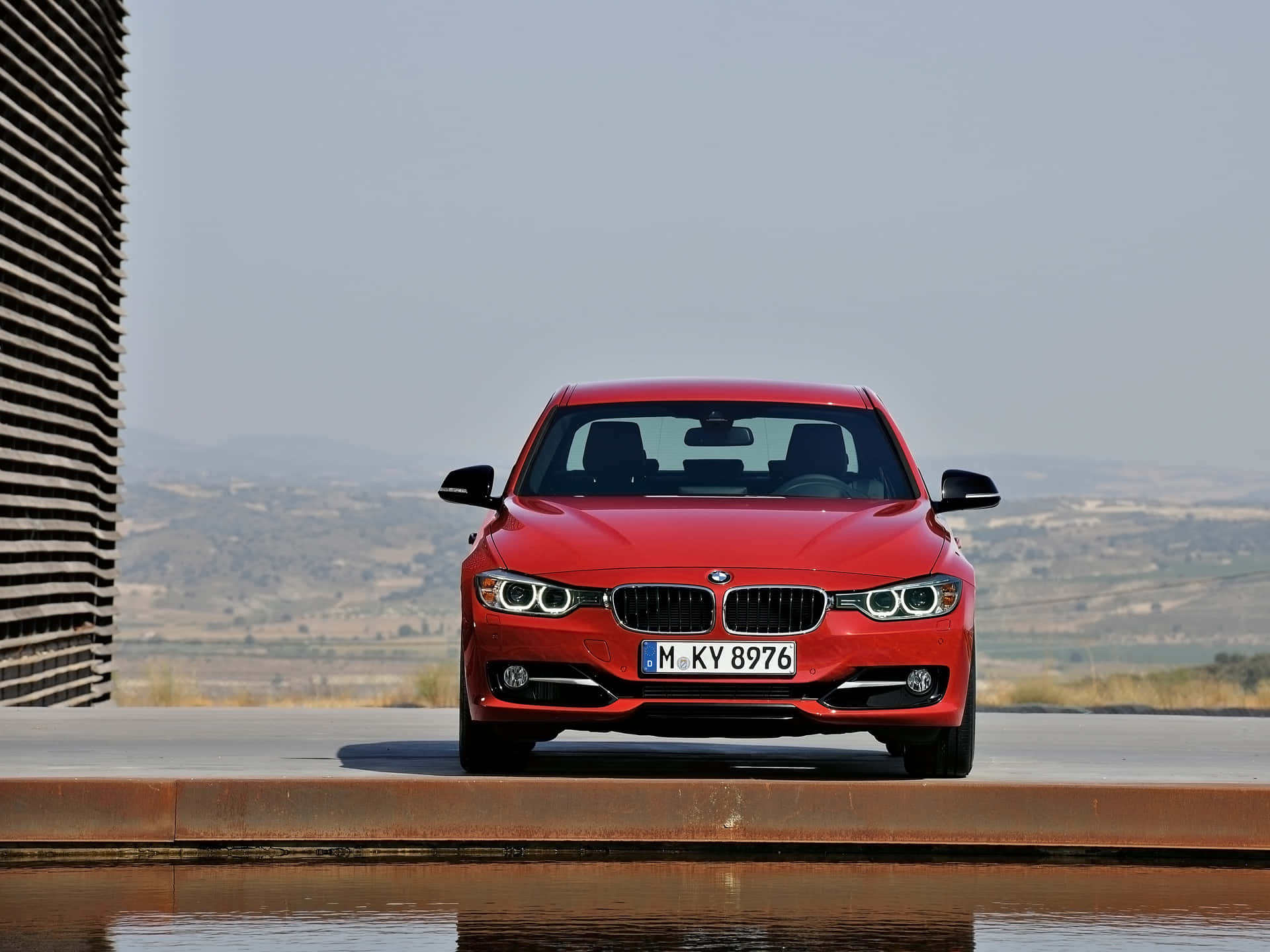 Bmw 328 桌布 壁紙 — 一輛鮮紅色的BMW轎車在混凝土平台上向前行駛，旁邊有水，背景有現代條紋牆和朦朧的山丘。